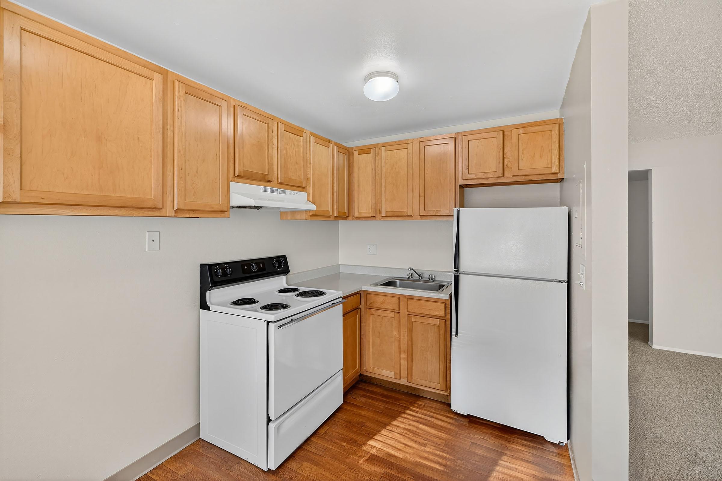 A small kitchen featuring light wooden cabinetry, a white stove with an oven, a white refrigerator, and a sink. The walls are painted light colors, and the flooring is a warm wood tone. There is a ceiling light providing illumination, and a doorway leading to another room is visible in the background.