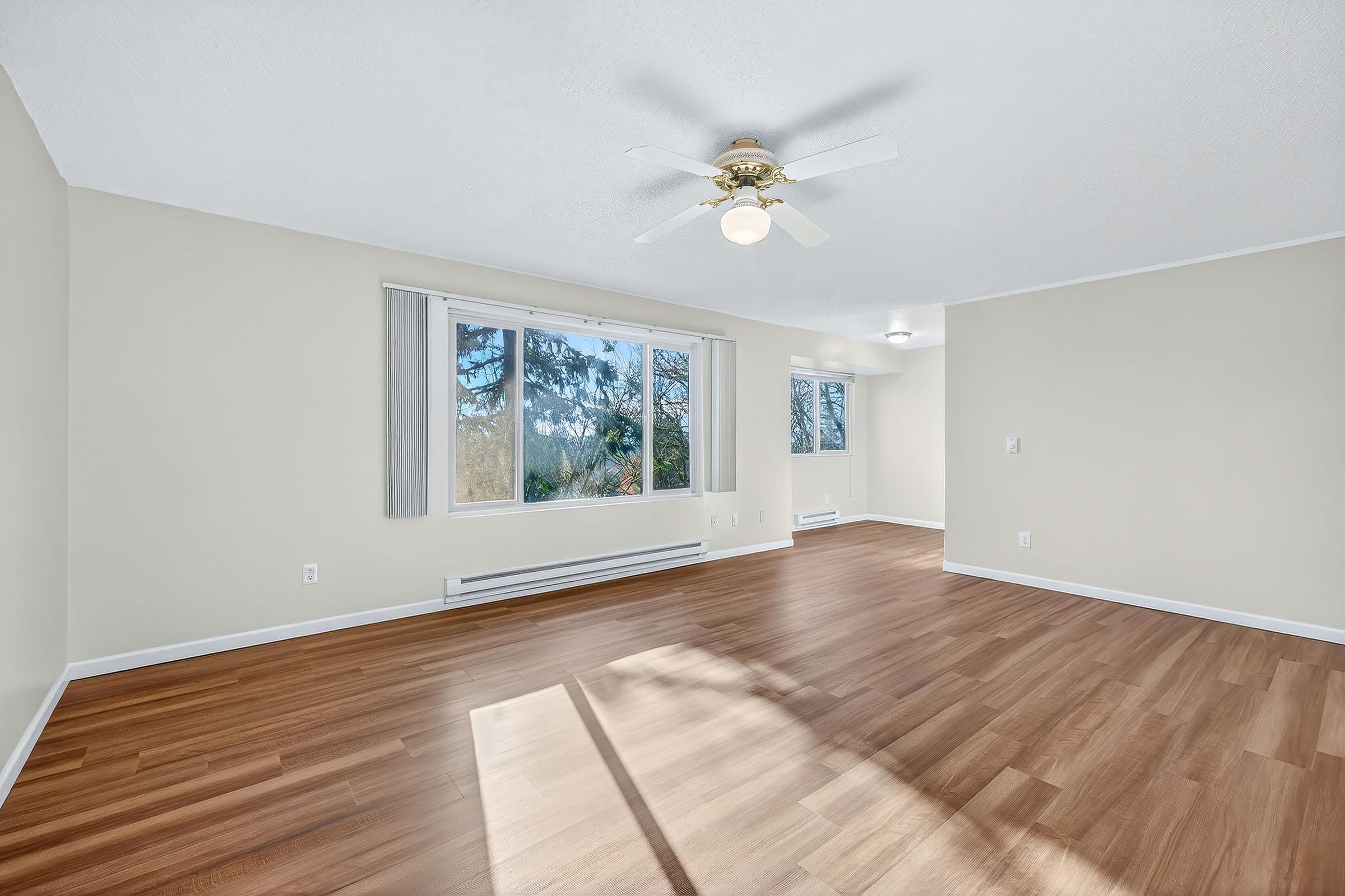 Spacious, well-lit interior of a room featuring a ceiling fan, large windows with a view of trees, and a modern wood floor. The walls are painted light, creating a bright and inviting atmosphere. There are baseboard heaters along the walls, and the room is devoid of furniture, offering a blank canvas for personalization.
