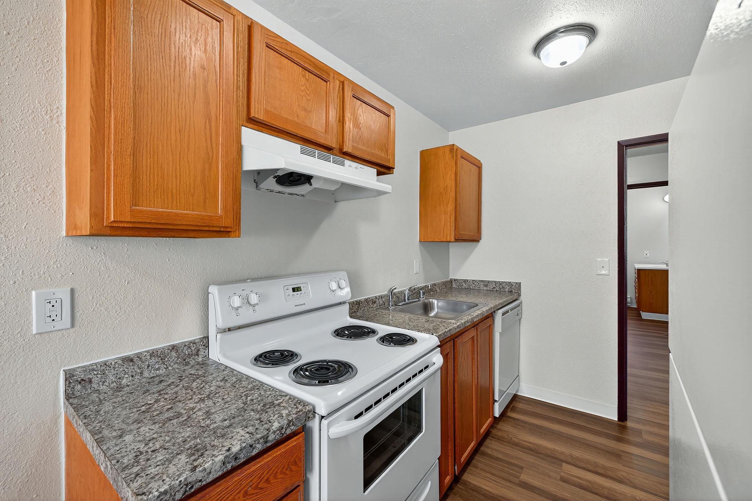 A modern kitchen featuring light wooden cabinets, a gray countertop, a white electric stove, an oven, and a hood. There is a single sink with a faucet and a door leading to another room in the background. The flooring is dark wood, and the walls are light-colored. Bright overhead lighting illuminates the space.