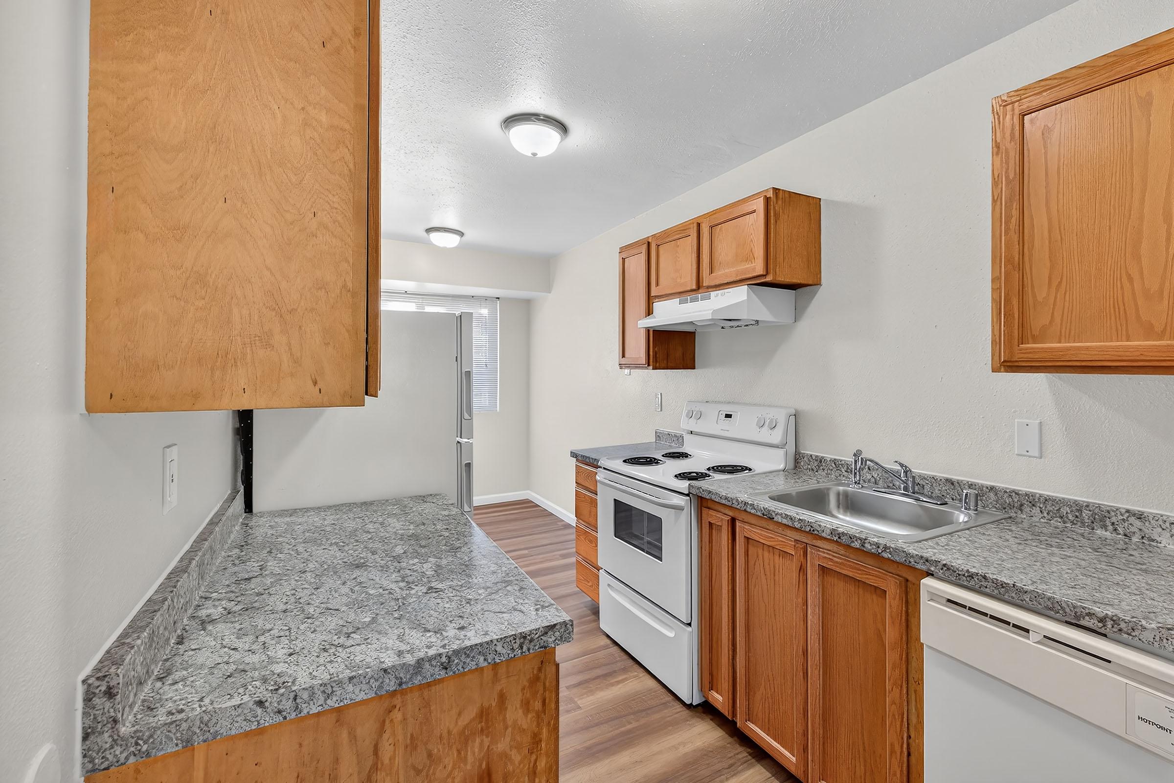 Bright and modern kitchen featuring wooden cabinetry, a granite countertop, a white stove, oven, and dishwasher. Natural light enters through a window, complemented by neutral-colored walls and wood-style flooring. The space is organized and functional, ideal for cooking and entertaining.