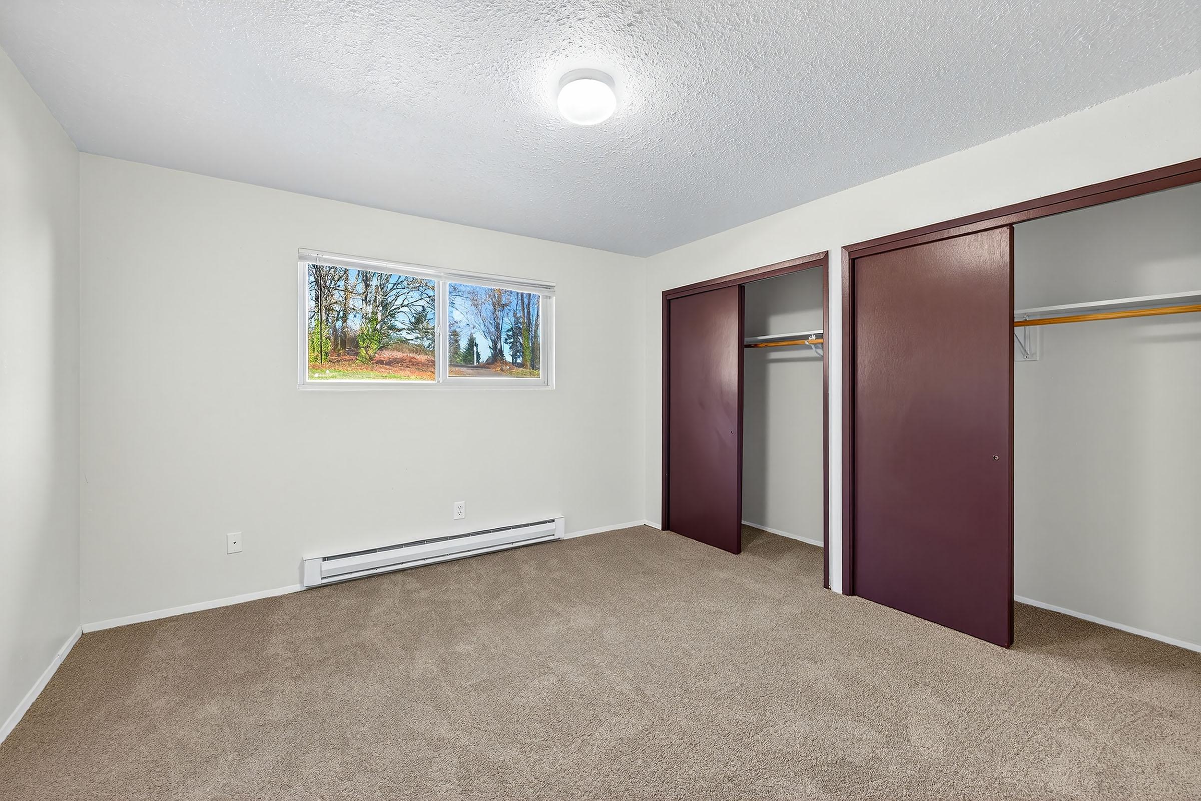 A vacant bedroom with beige carpeting and light-colored walls. It features a window with a view of trees outside, and two dark brown closet doors are open, revealing empty closet space. The room is well-lit by a ceiling light fixture.