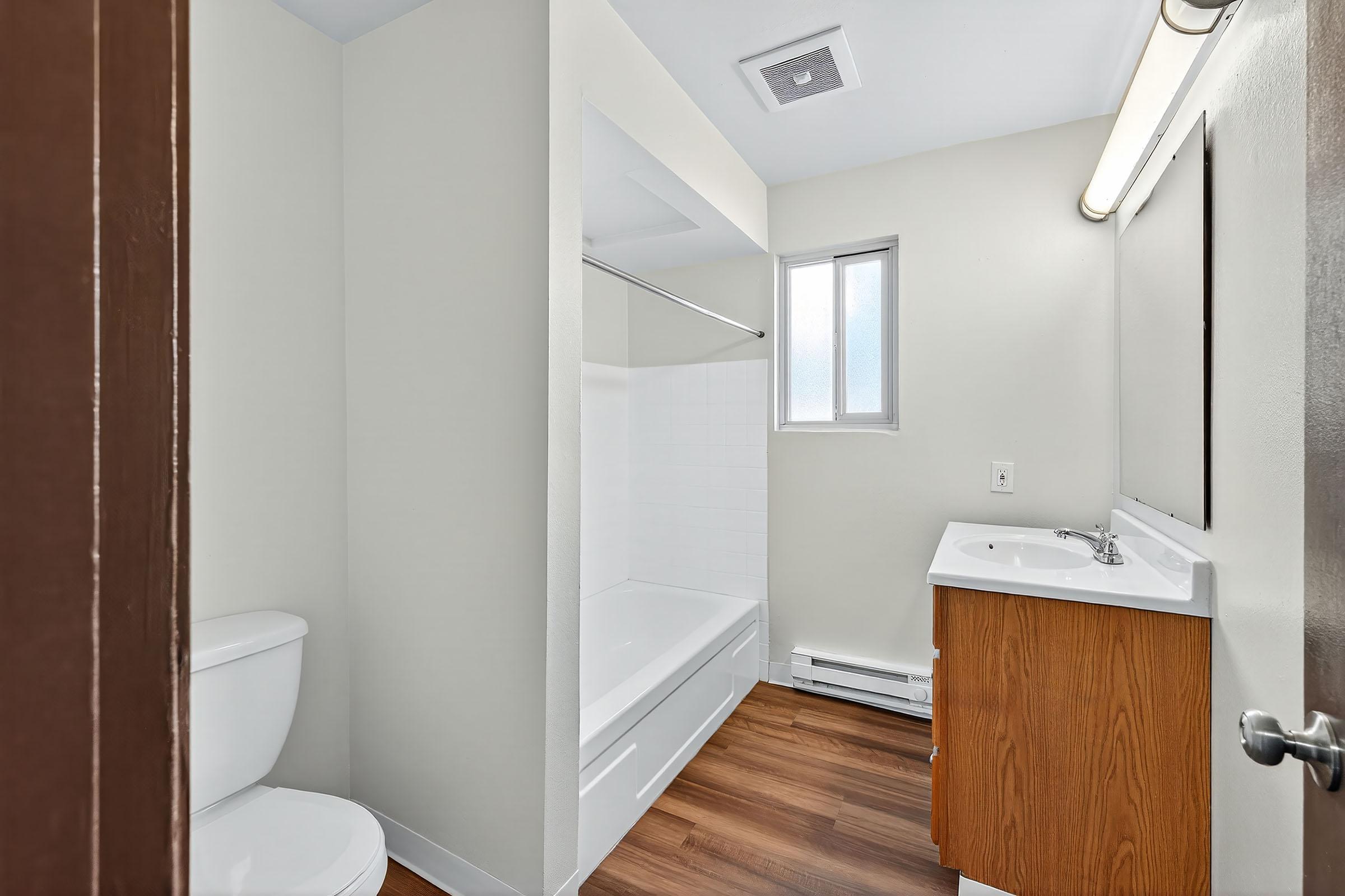 A clean and modern bathroom featuring a toilet, a bathtub with a shower, a sink with a wood vanity, and a window providing natural light. The walls are painted a light color, and the flooring is a warm wood finish.
