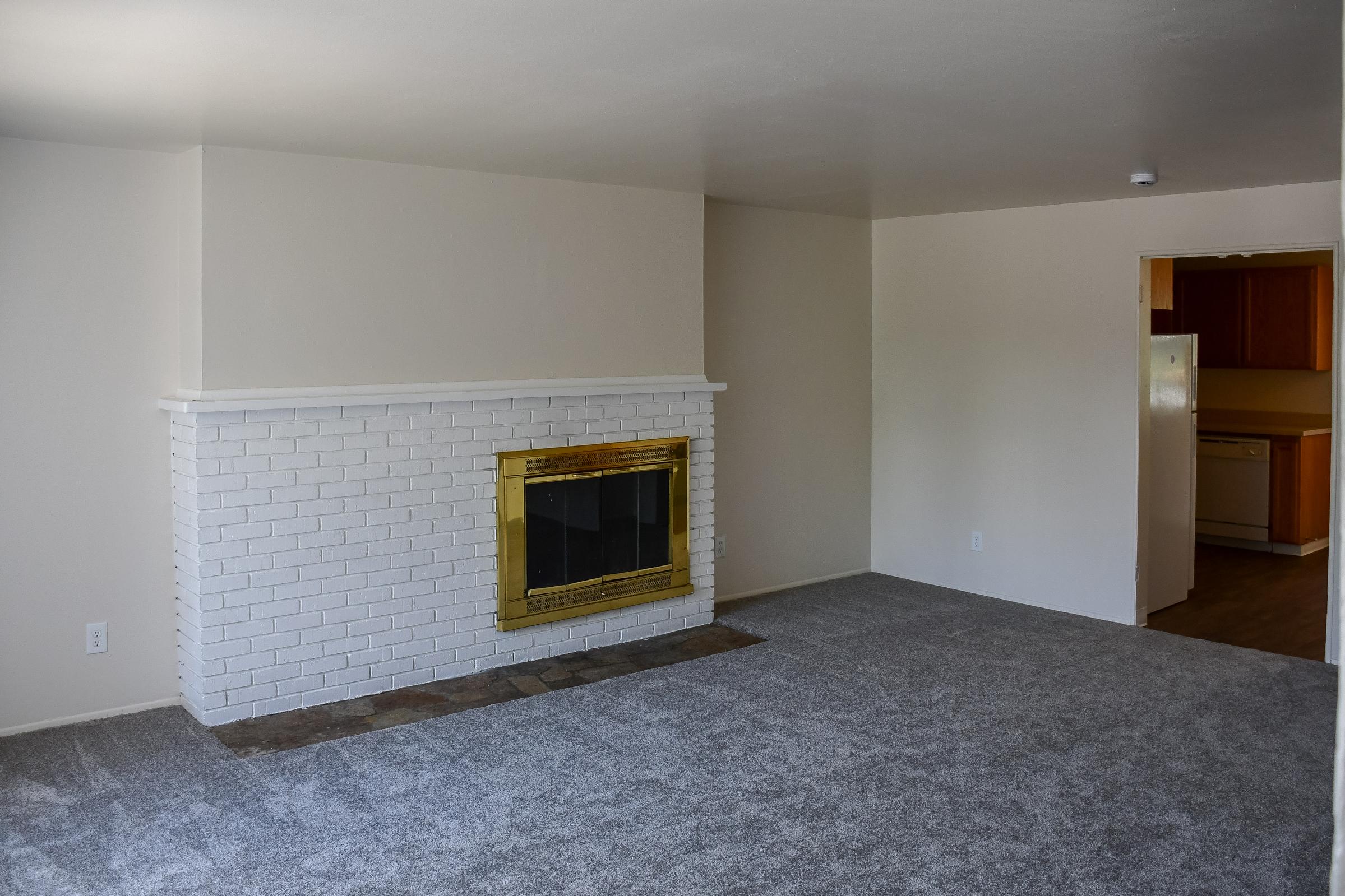 A spacious living room featuring a white brick fireplace with a brass trim, light gray carpet, and plain white walls. An open doorway leads to a kitchen with wooden cabinets. The room is well-lit and uncluttered, offering a minimalist feel.