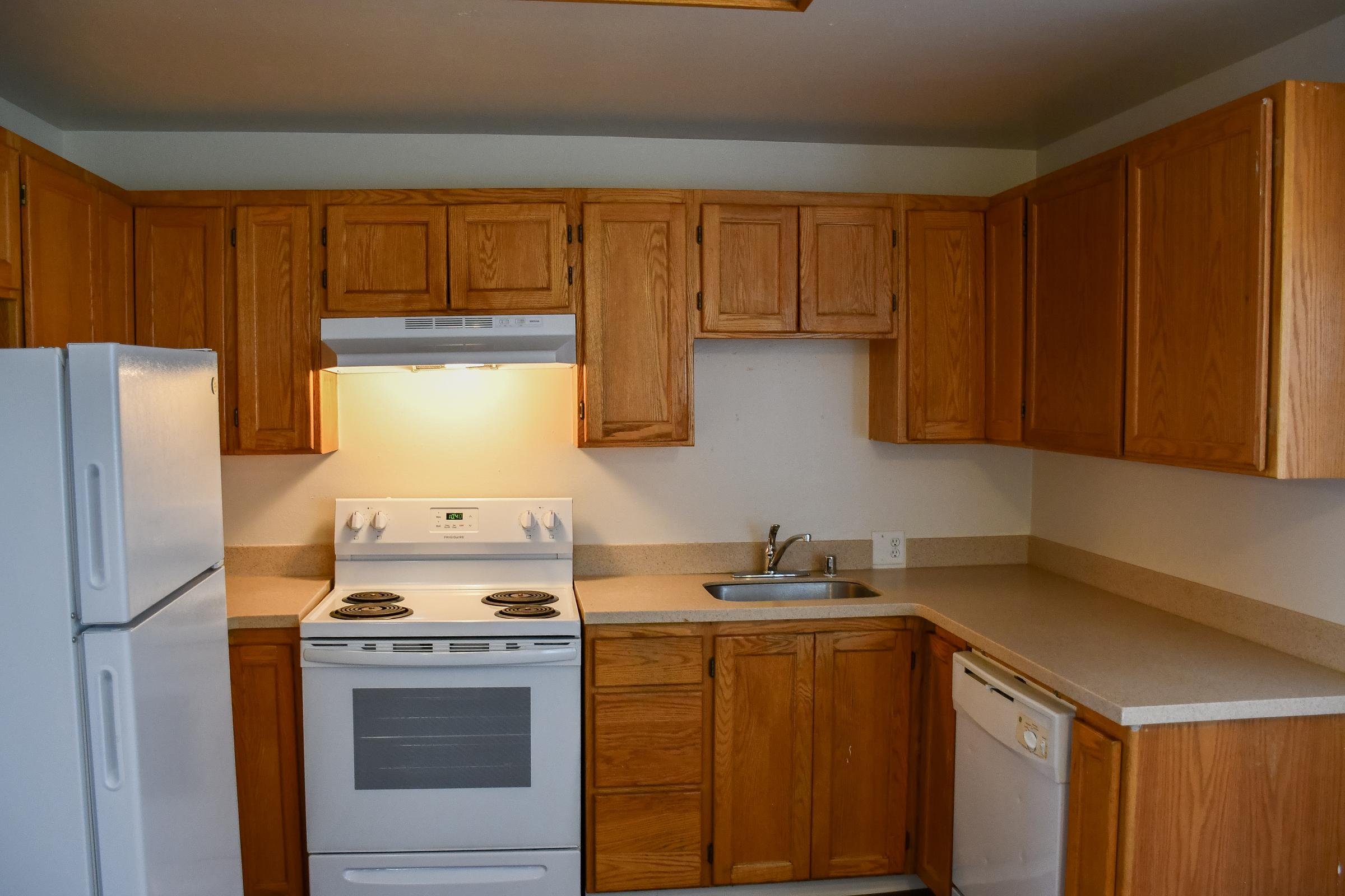 A compact kitchen featuring wooden cabinets, a white stove, a double sink, a refrigerator, and a dishwasher. The countertops are light-colored, and the kitchen has a neutral wall tone, creating a warm and functional space.
