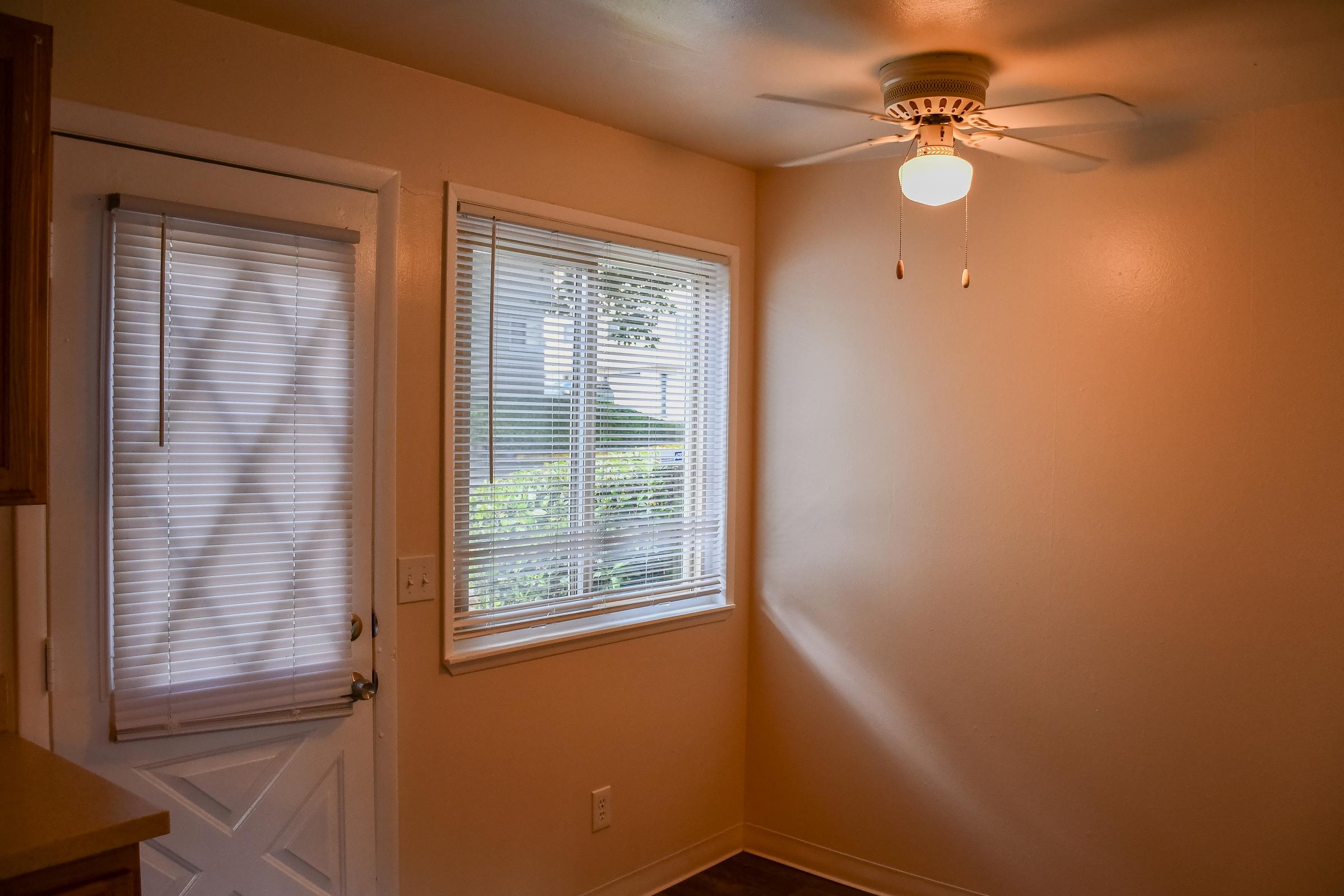 A well-lit room with peach-colored walls, featuring a ceiling fan and a window with blinds. There’s a door to the left and a simple light fixture overhead. The space appears clean and inviting, with natural light filtering through the window, illuminating the room's modest decor.