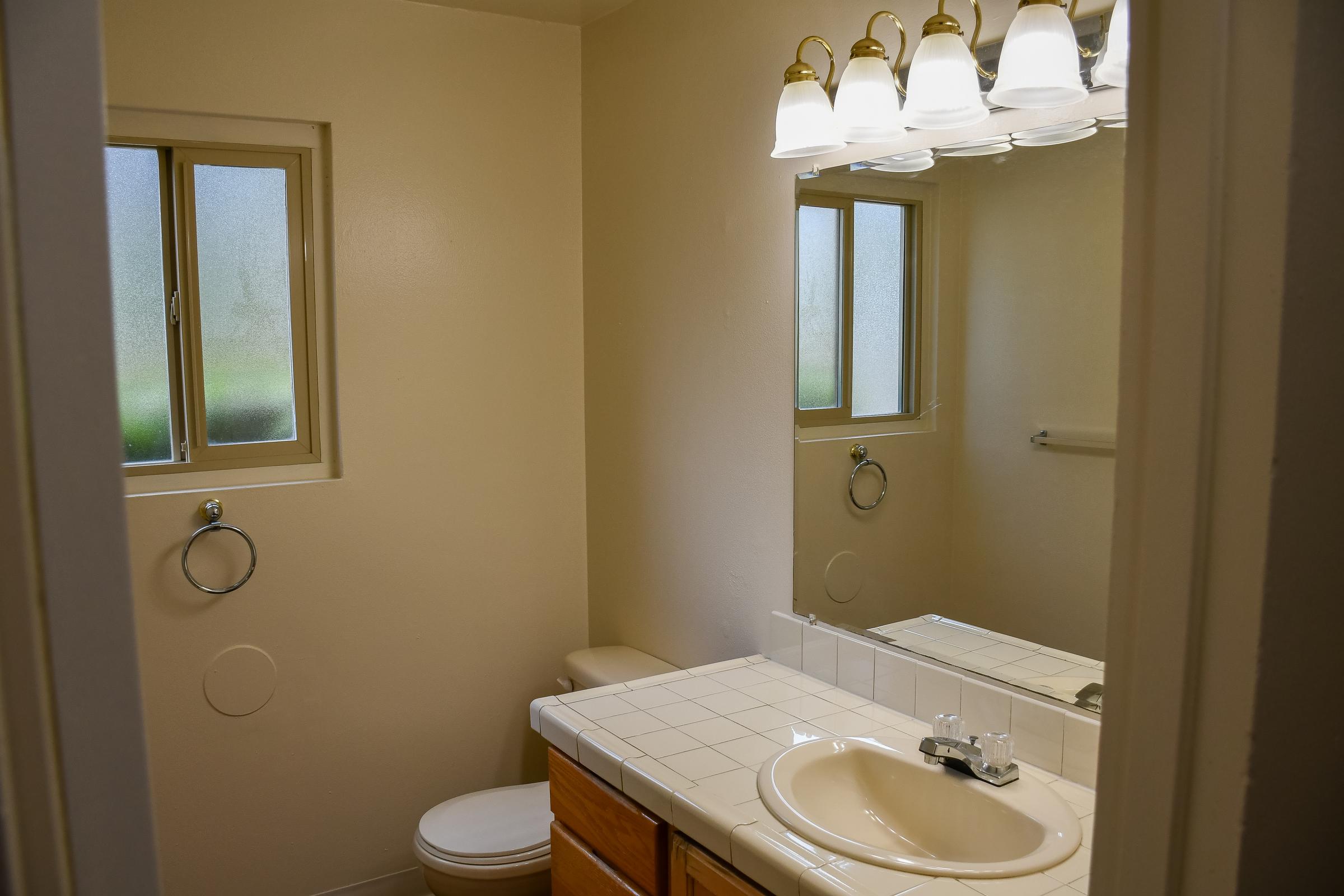 A small bathroom featuring a white sink with a faucet, a beige countertop, a toilet, and a mirror above the sink. The room has two frosted glass windows allowing natural light, and a light fixture with multiple bulbs is mounted above the mirror. The walls are plain and painted in a light color.