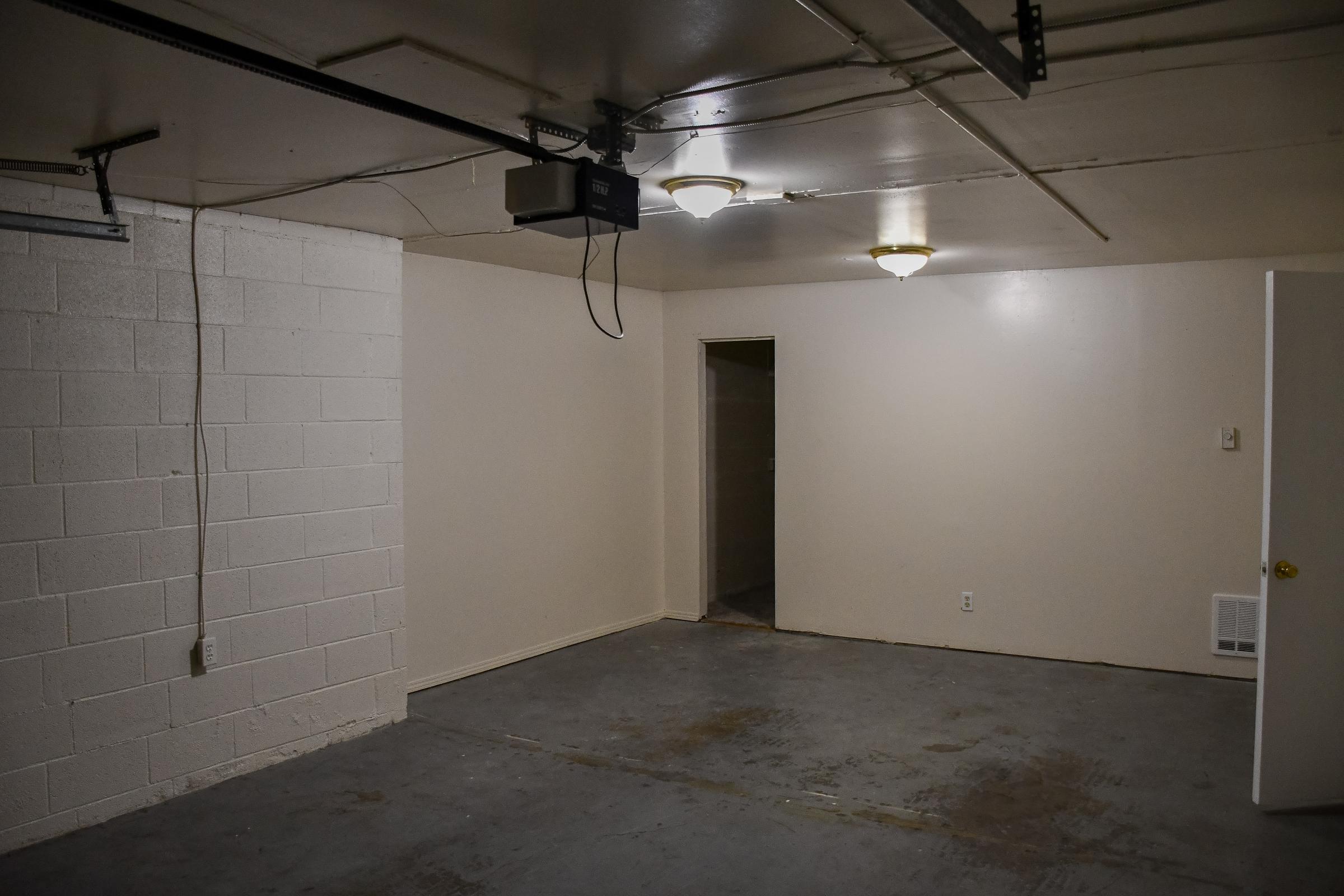 An empty, well-lit room with white cinder block walls and a concrete floor. There are two ceiling lights, a door on the right leading to another space, and a wall-mounted projector near the ceiling. The room appears to be clean and sparsely furnished.