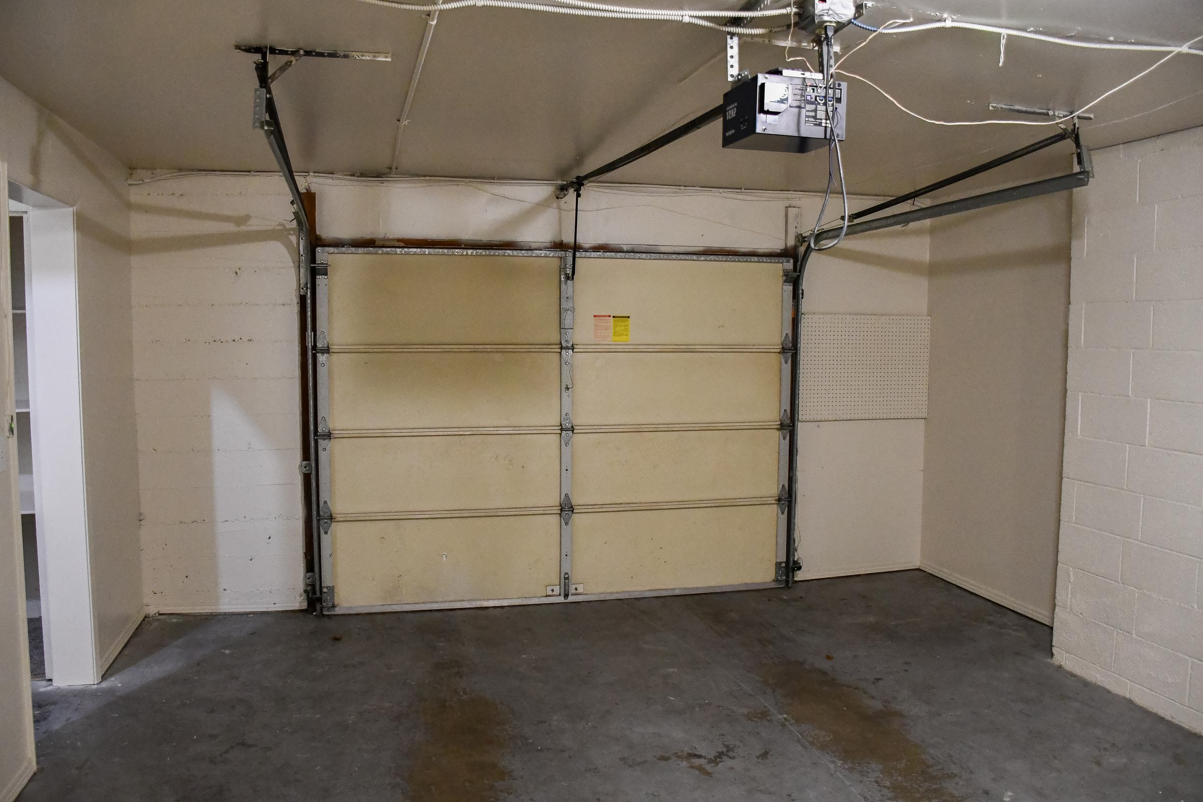 Interior of a garage featuring a large garage door, concrete flooring, and plain white walls. A garage door opener is mounted on the ceiling, and there is a small door on the left side leading to another space. The environment appears clean and empty, suitable for storage or parking.