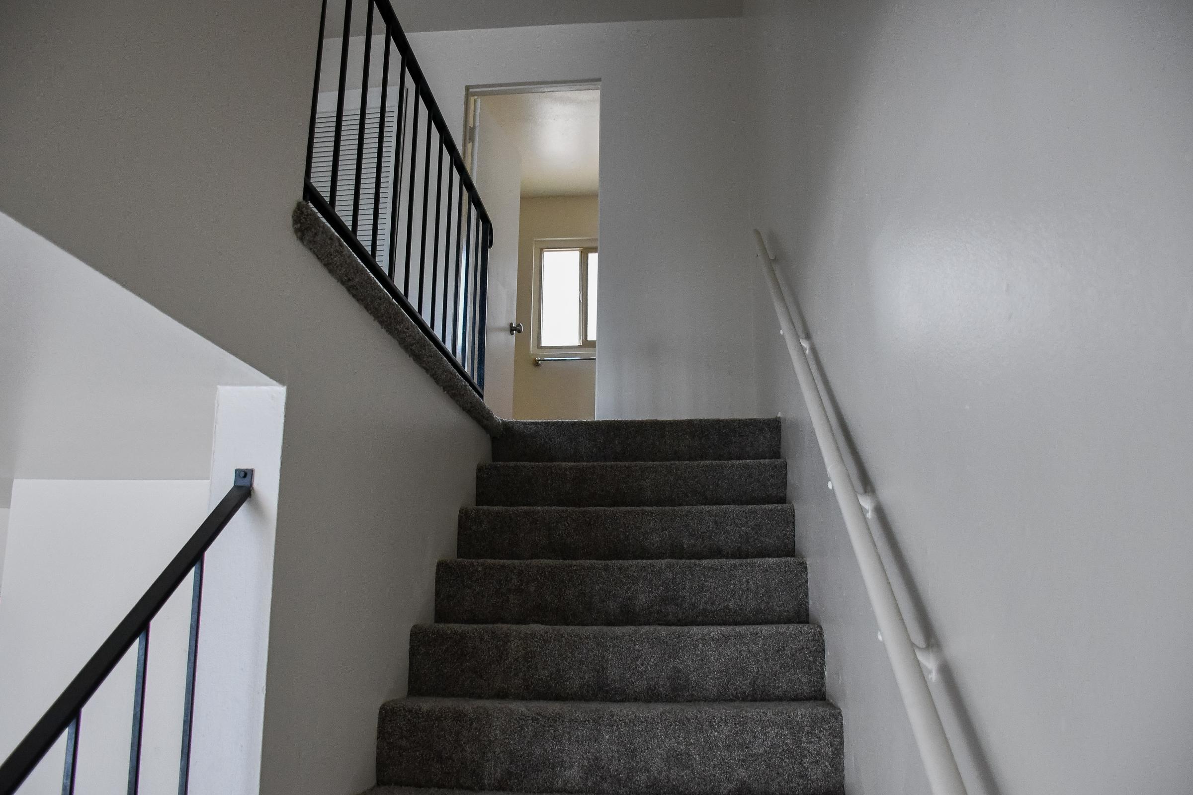Staircase leading up with gray carpet, a metal railing, and a white wall. At the top of the stairs, a doorway can be seen with a window beside it, allowing natural light to enter the space.
