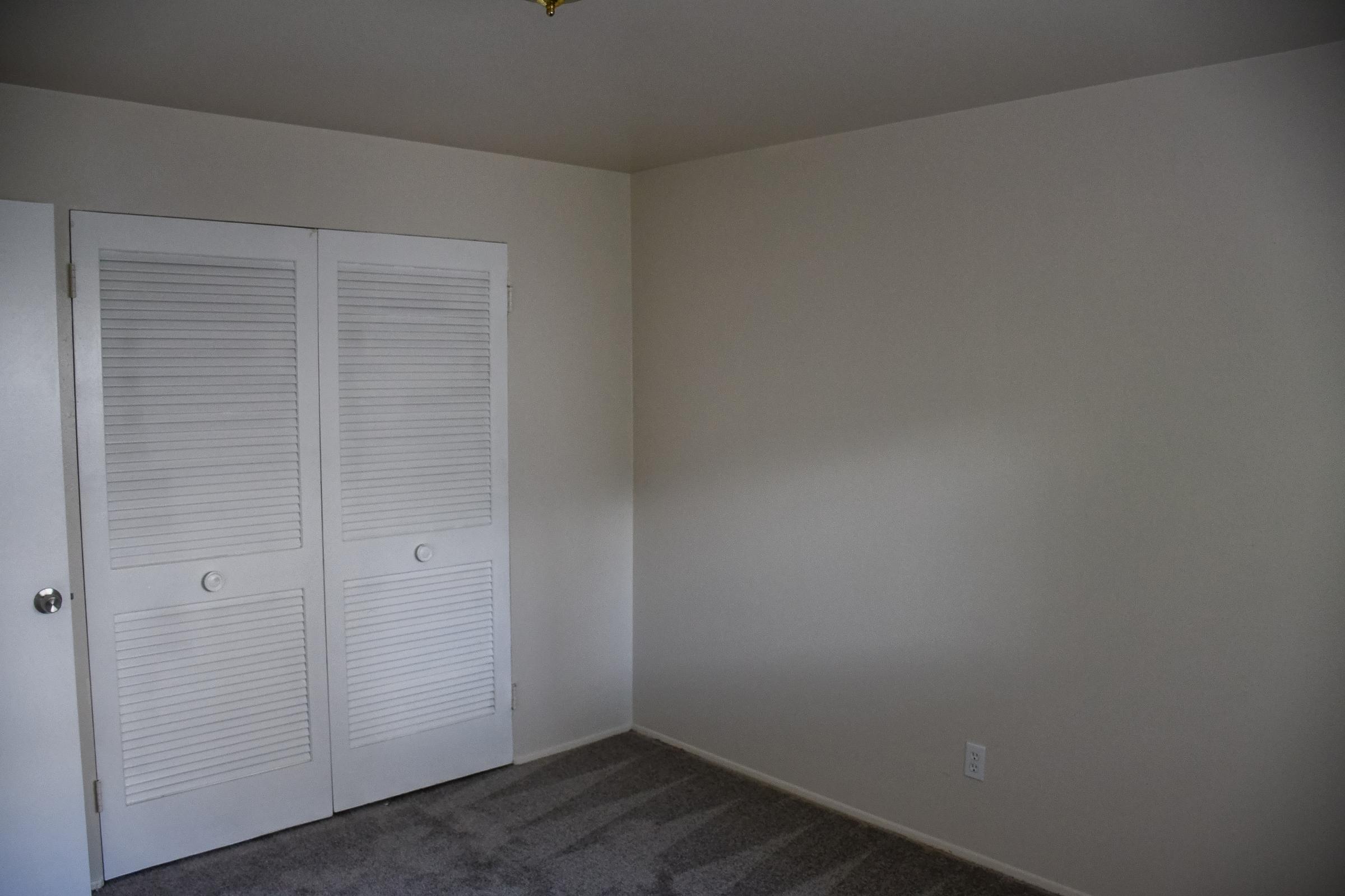 A minimally furnished room featuring a light-colored wall, carpeted floor, and a set of white double doors with louvered panels. The space is unadorned and well-lit, with no furniture or decorations present.