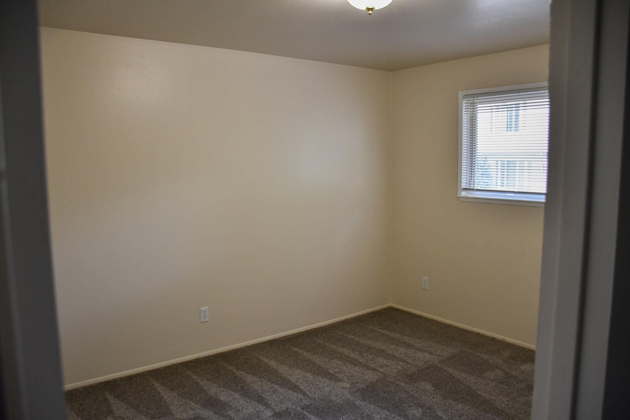 A vacant room featuring light beige walls and gray carpet. The space includes a single window with white blinds, allowing natural light to enter. The ceiling has a light fixture, and there are no furnishings in the room. The overall appearance is clean and simple, suitable for various uses.