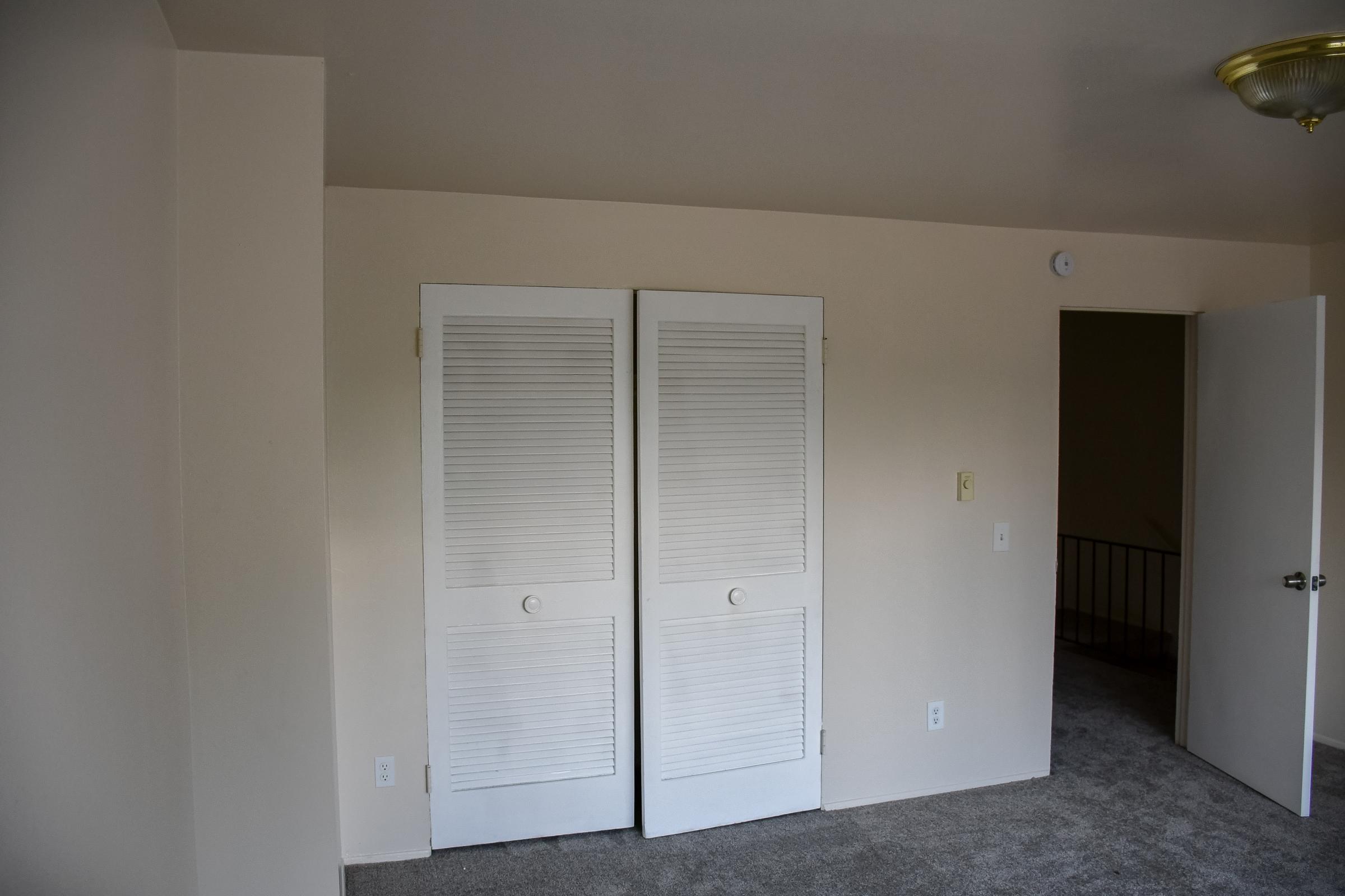 An empty room featuring beige walls and a light-colored carpet. There are double white louvered doors on the left side leading to a closet, and a door on the right side that opens to another area. A ceiling light fixture is visible above, and the room appears bright and uncluttered.