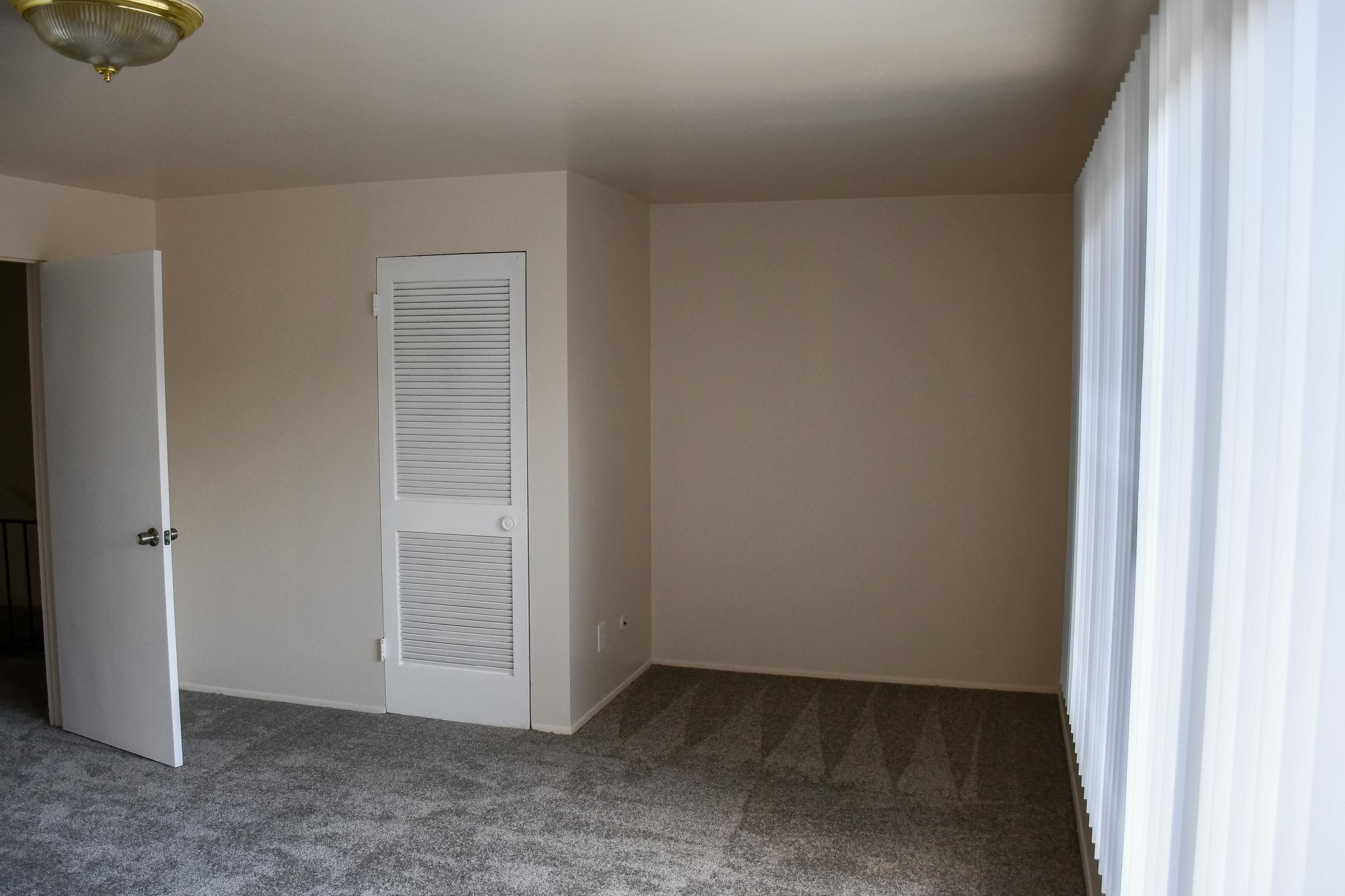 A spacious, empty room featuring light-colored walls and carpet. A closed door with a louvered design is located on the left, while large windows with sheer curtains allow natural light to fill the space. The ceiling has a simple circular light fixture, creating a bright and airy atmosphere.