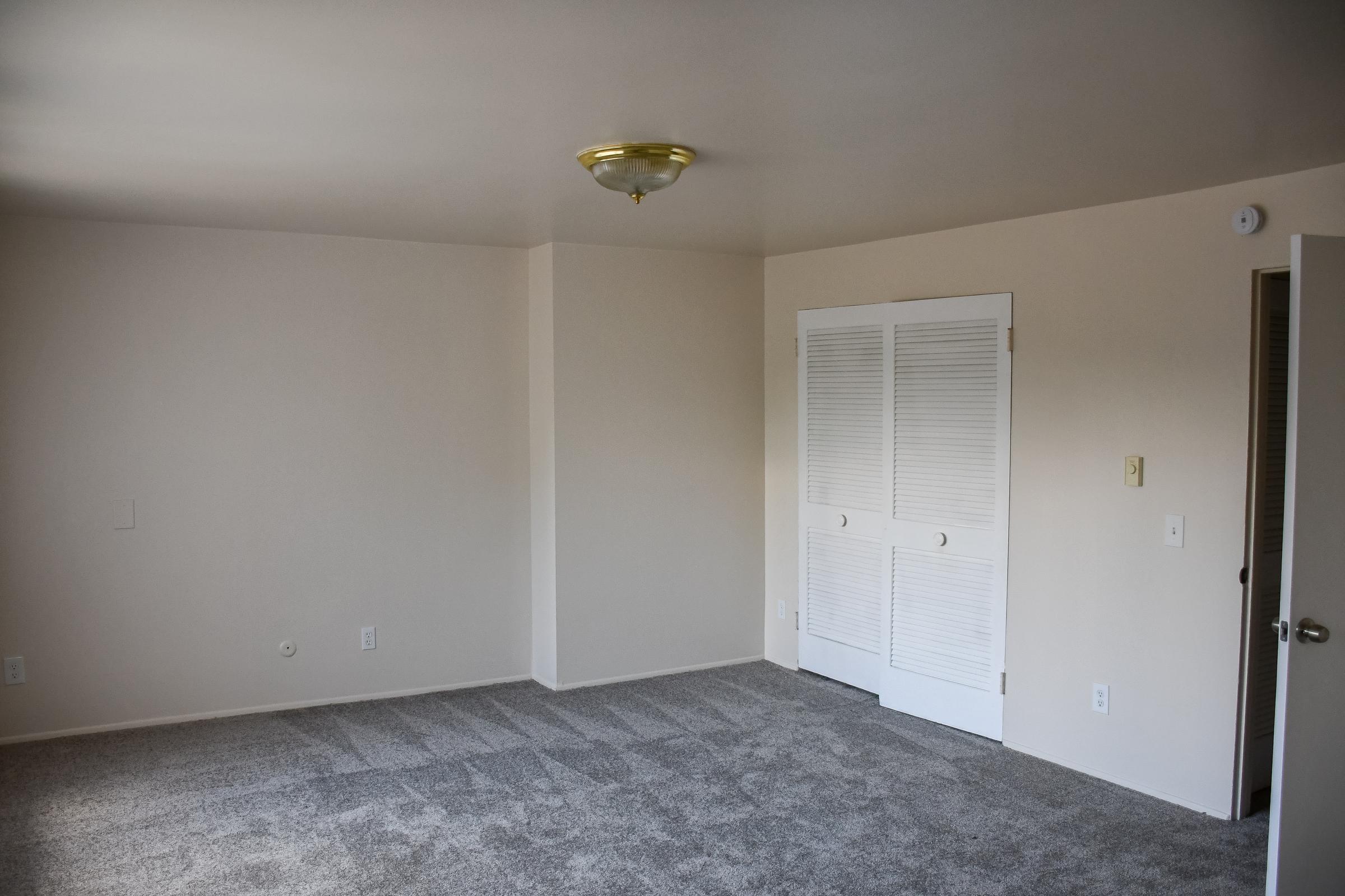 A well-lit, empty room with beige walls and a light-colored carpet. The room features a ceiling light fixture and a closed white double door closet. There are two plain wall outlets visible. The overall atmosphere is clean and neutral, ideal for personal customization.