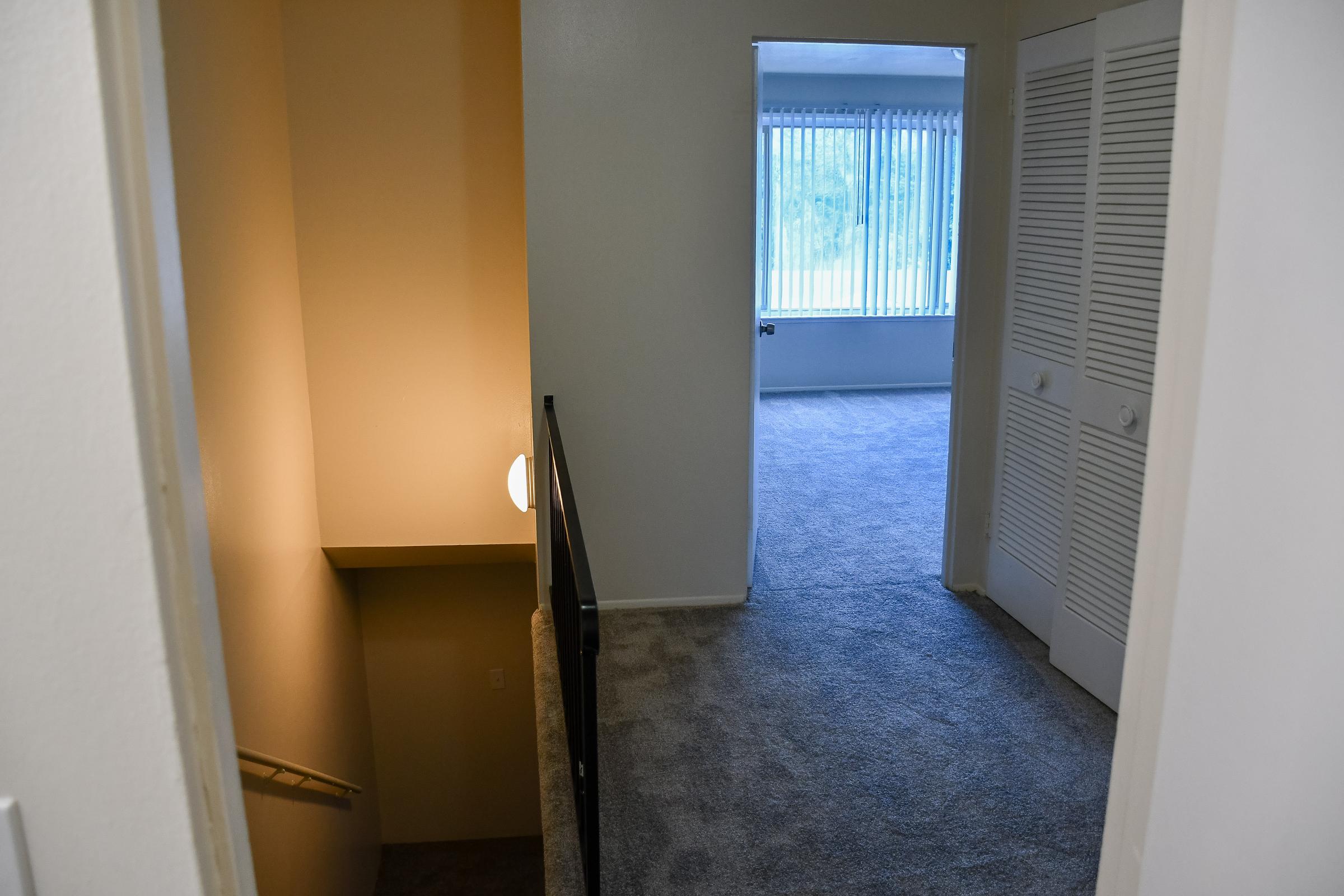 A view of an interior hallway overlooking a staircase leading down, with a light fixture illuminating the space. To the right, there's an open room featuring large windows with vertical blinds and carpeted floors, while a closed closet with sliding doors is visible on the left side.