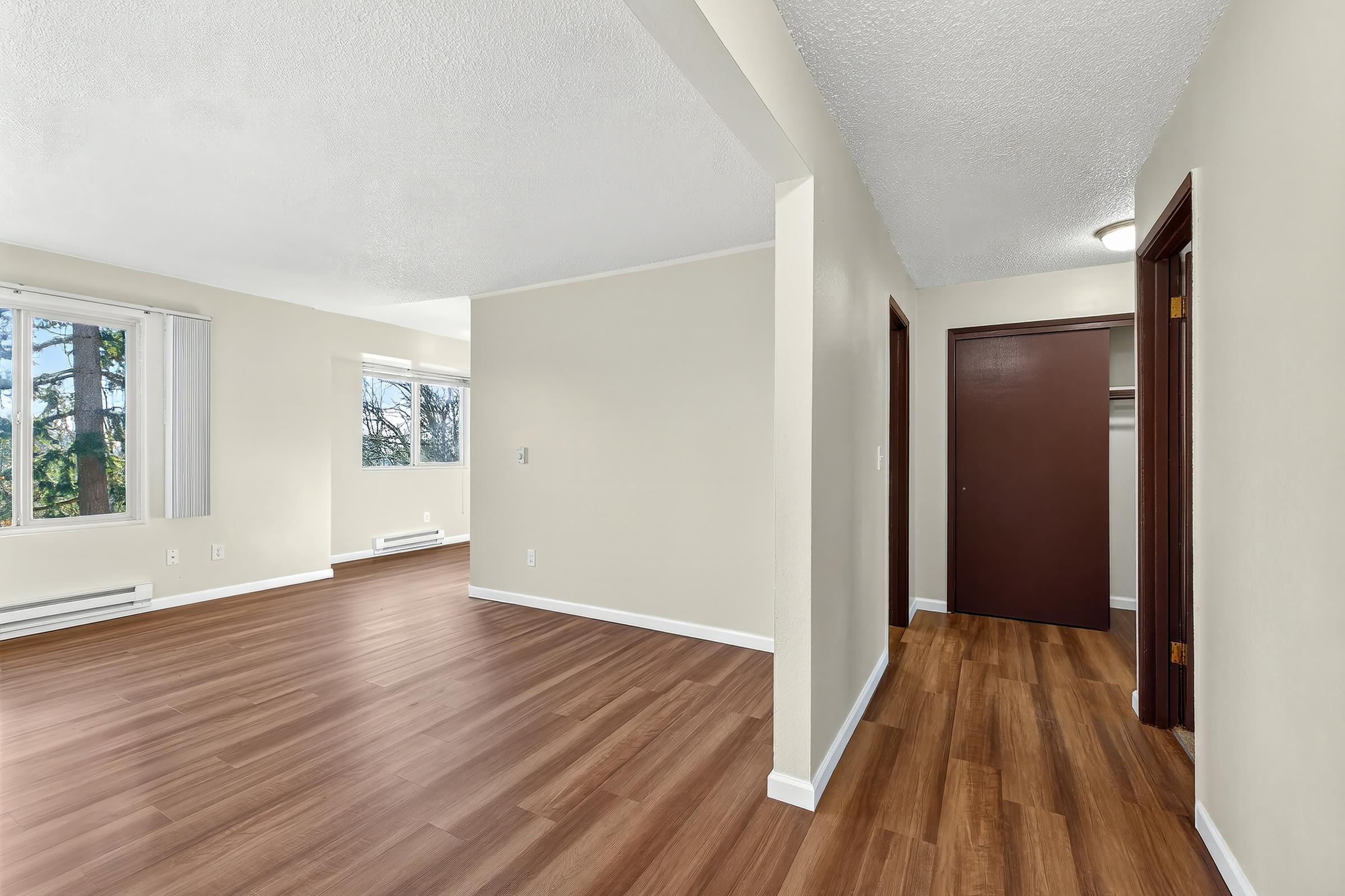 An interior view of a spacious apartment with light-colored walls and wood laminate flooring. The image shows a living area on the left with large windows allowing natural light, and a hallway leading to a closed door on the right, indicating a closet or room.