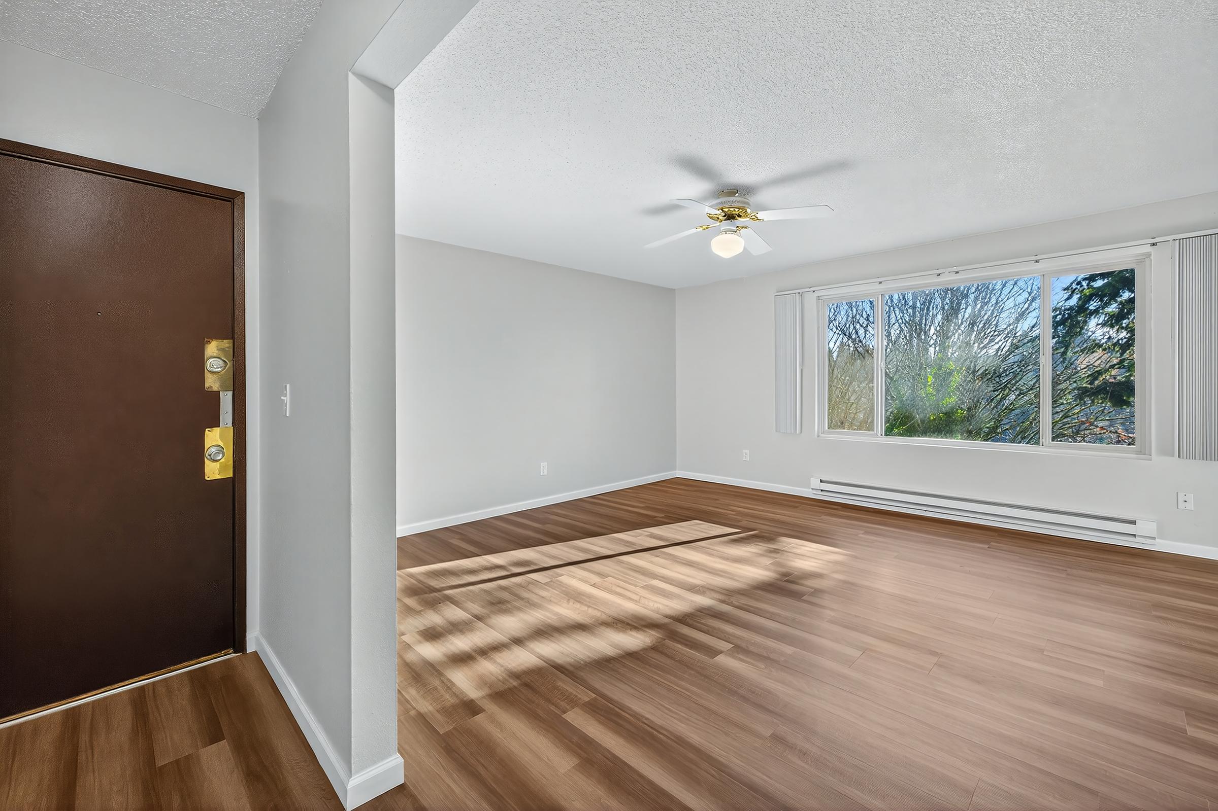 A spacious living room featuring a large window with natural light, light hardwood flooring, and a ceiling fan. The entrance door is visible on the left, and the walls are painted in a neutral color, creating a bright and inviting atmosphere.
