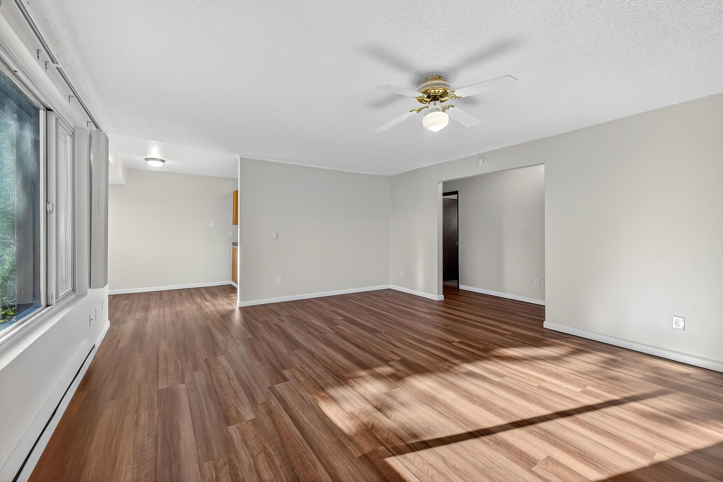 A spacious, well-lit living room featuring light-colored walls, hardwood flooring, and a ceiling fan. Large windows provide natural light, while an open layout leads to adjacent rooms, including a kitchen visible through an archway.
