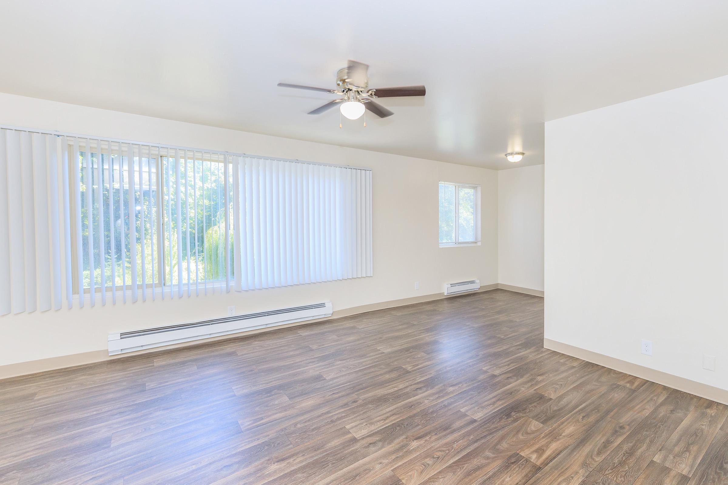 Spacious, empty room with light-colored walls and laminate flooring. Large windows with vertical blinds let in natural light, and a ceiling fan is visible. There are heating vents along the walls, and the overall ambiance is bright and airy, suitable for various interior design options.