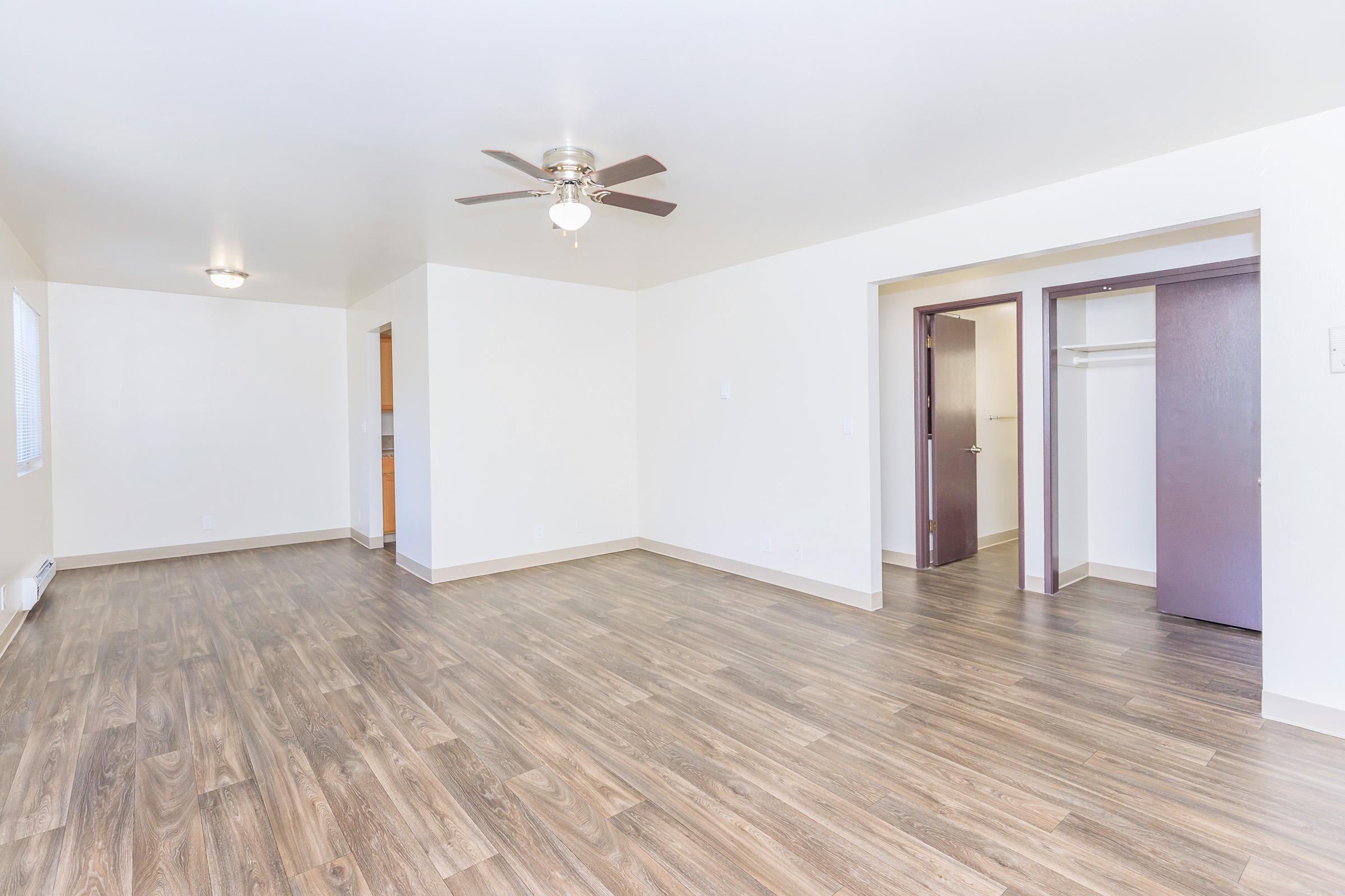 Spacious interior of an empty room featuring wooden flooring, a ceiling fan, and neutral-colored walls. There are doorways leading to a closet and a kitchen area, with plenty of natural light coming through the window. The overall design is modern and inviting, ideal for furnishing and personalizing.