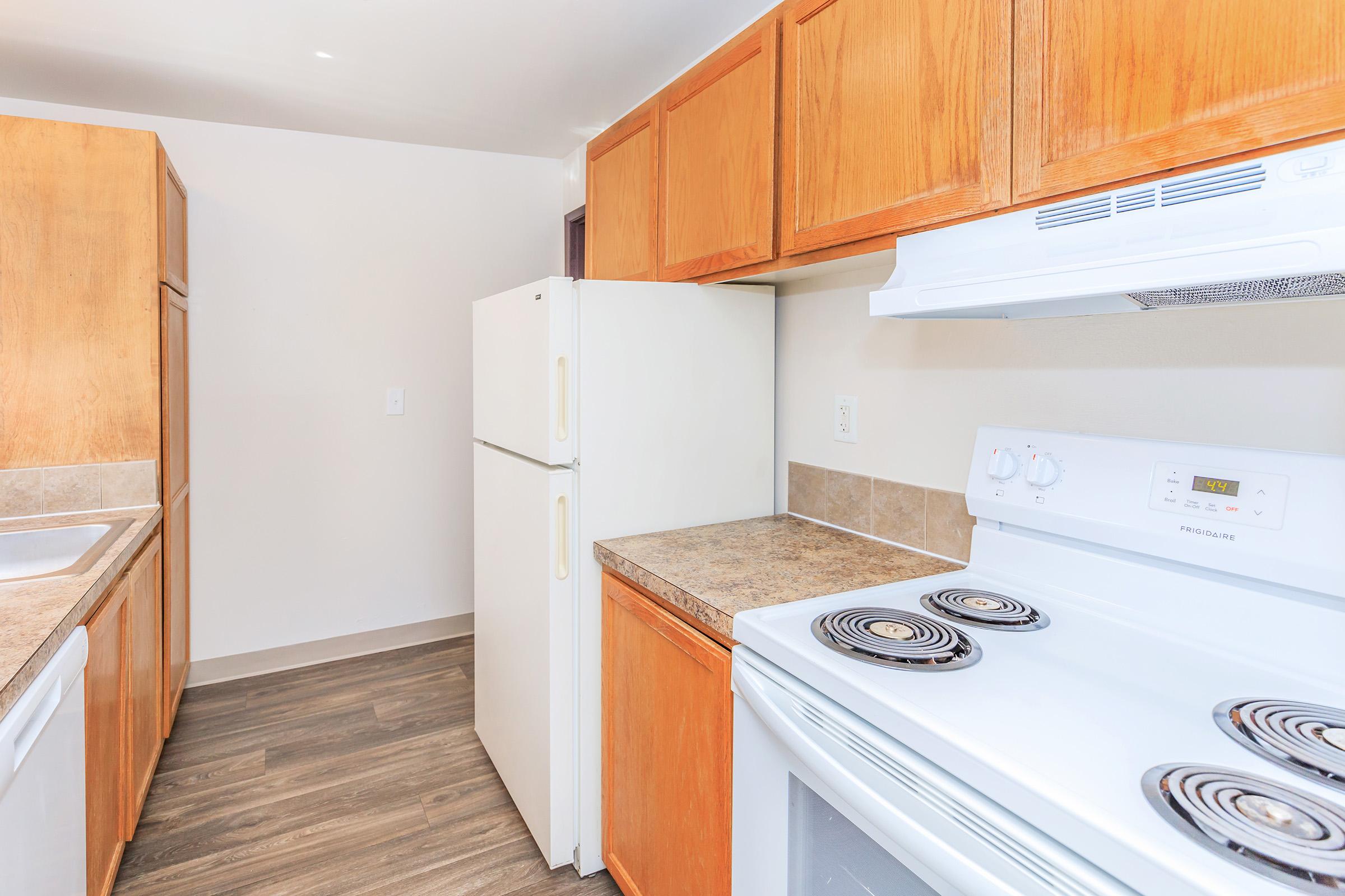 A kitchen interior featuring wooden cabinets, a white stove, and a refrigerator. The countertops are light-colored, and the flooring is a wood-like laminate. The room has good natural light and a simple layout, creating a clean and functional cooking space.