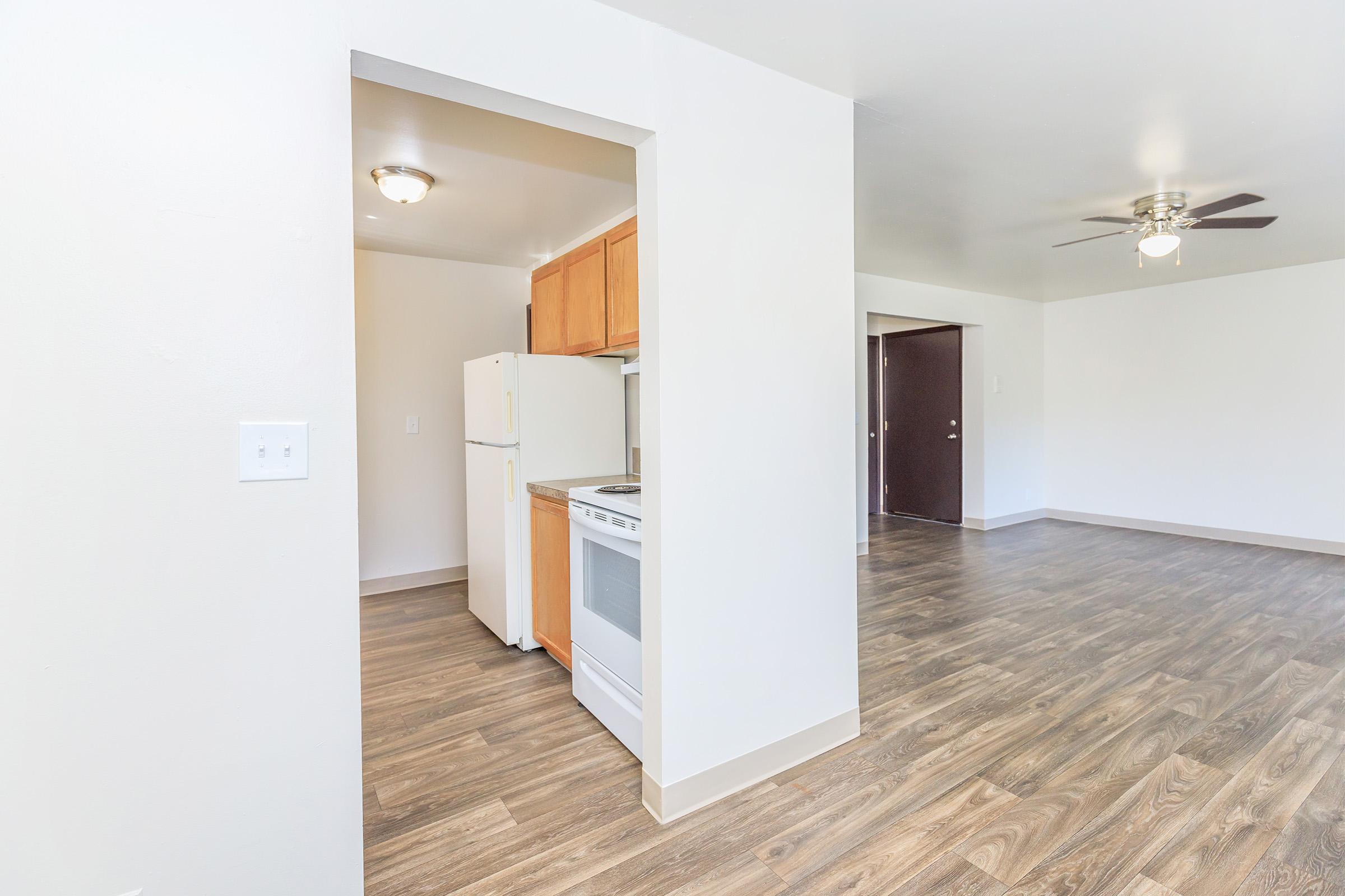 Interior view of a room showing an open layout. To the left, there is a kitchen area with a white refrigerator and stove. The floor is wood laminate, and the walls are painted in a light color. In the background, there are two doorways leading to another room and an entrance, with a ceiling fan visible.