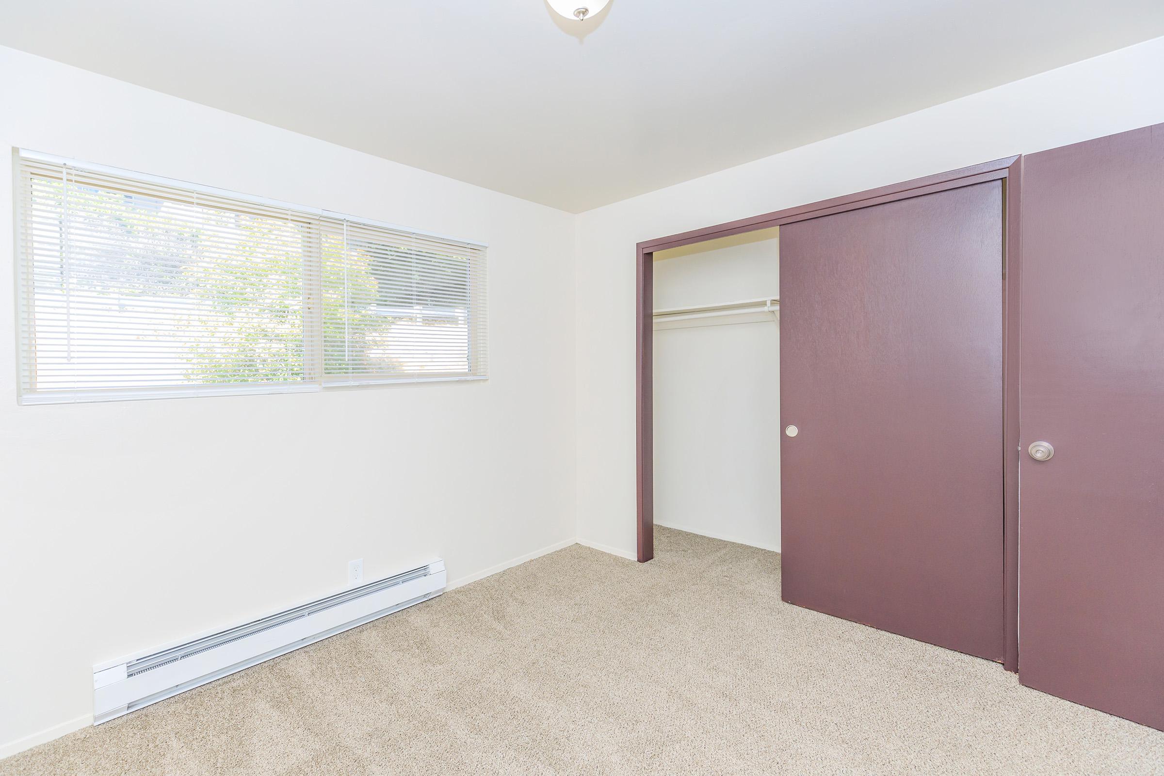 A bright, empty room with beige carpet and white walls. It features a window with blinds allowing natural light, a sliding closet door in a rich purple color, and a baseboard heater along one wall. The overall atmosphere is clean and ready for personalization.