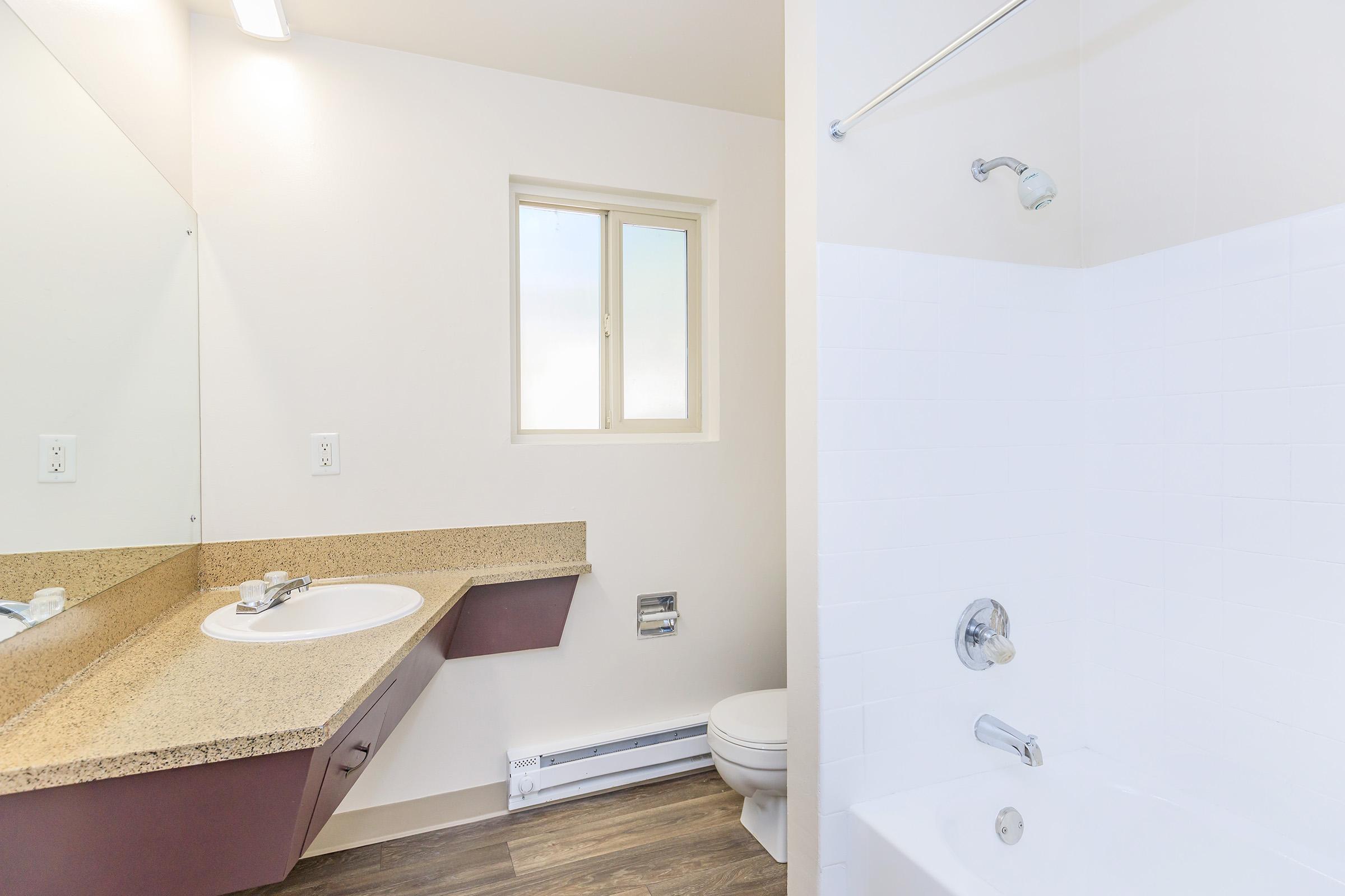A clean, modern bathroom featuring a white bathtub with a showerhead, a small window letting in natural light, and a countertop with a sink. The walls are light-colored, and there is a single mirror above the sink. The floor is made of wood-like material, enhancing the contemporary look.
