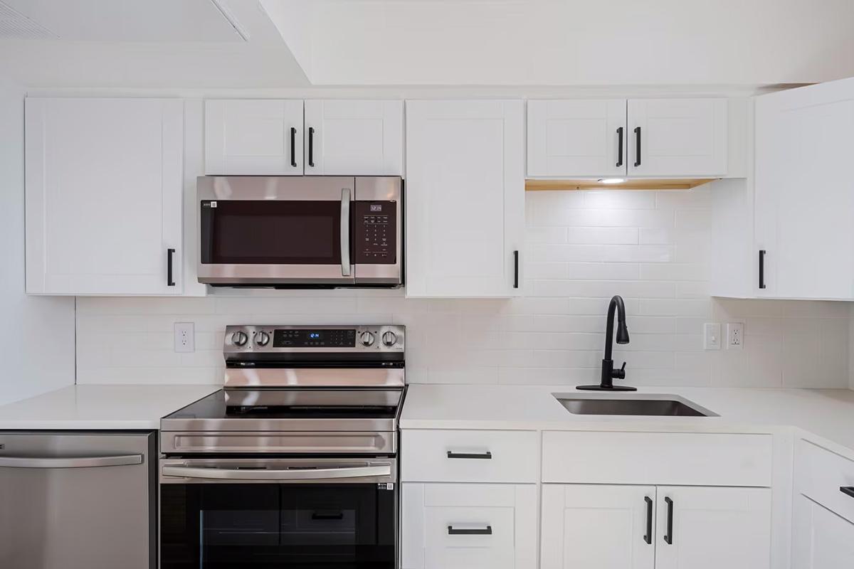 Modern kitchen featuring white cabinetry, stainless steel appliances including a microwave, oven, and refrigerator. A black faucet is installed above a single bowl sink, with a subtle backsplash and under-cabinet lighting providing a clean, contemporary look.