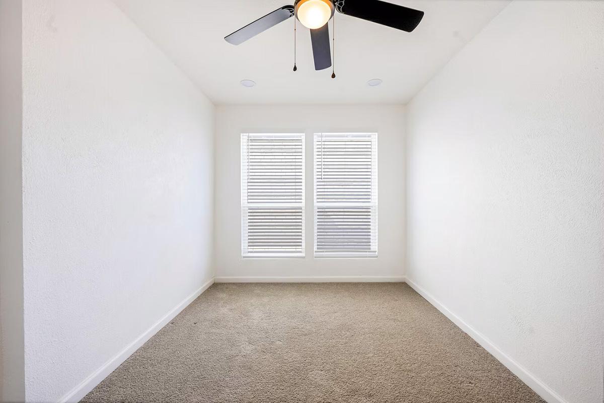 A small, empty room with light-colored walls and carpeted flooring. It features two windows with white blinds, allowing natural light to enter. A ceiling fan is installed, and the overall space appears clean and minimalistic.