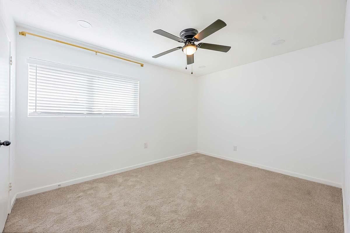 Empty room with light-colored walls and beige carpet. A ceiling fan hangs from the ceiling, and a window with white blinds allows natural light to enter. The room has a simple, uncluttered design, making it suitable for various purposes.