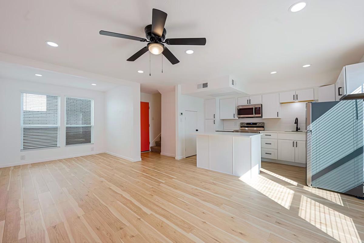 Modern open-concept living space featuring light wood flooring, a ceiling fan, white walls, and large windows. The kitchen includes stainless steel appliances and a central island, with a glimpse of a staircase in the background. Natural light brightens the area, creating a welcoming atmosphere.