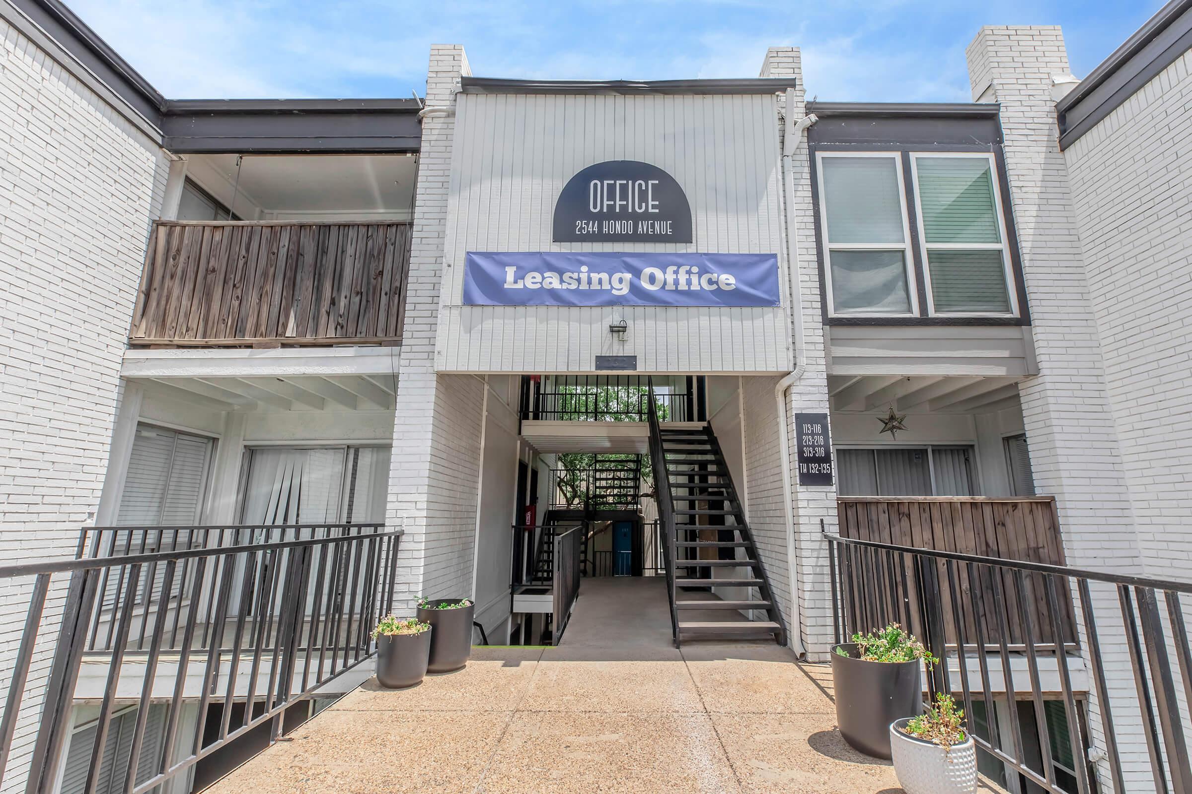 Leasing office entrance with a sign that reads "Office" above and "Leasing Office" prominently displayed on a blue banner. The building features white brick, balconies with wooden railings, and potted plants flanking the pathway leading to the entrance.