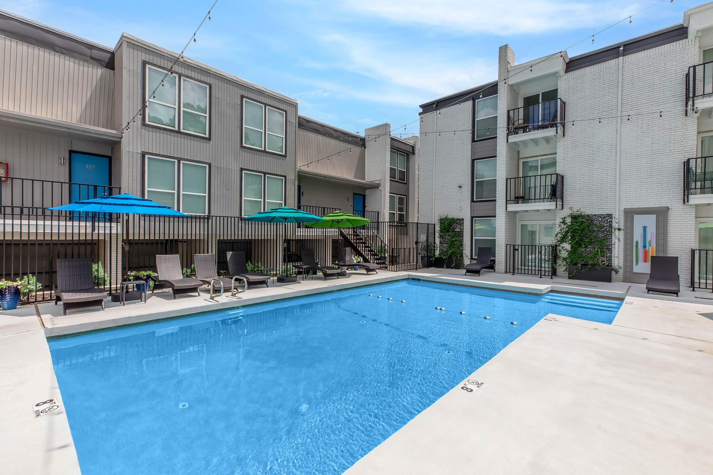 A clear blue swimming pool surrounded by lounge chairs and umbrellas in a modern apartment complex courtyard. The area features stylish architecture with a mix of light and dark-colored buildings, and plants adding greenery. The atmosphere is inviting and perfect for relaxation.