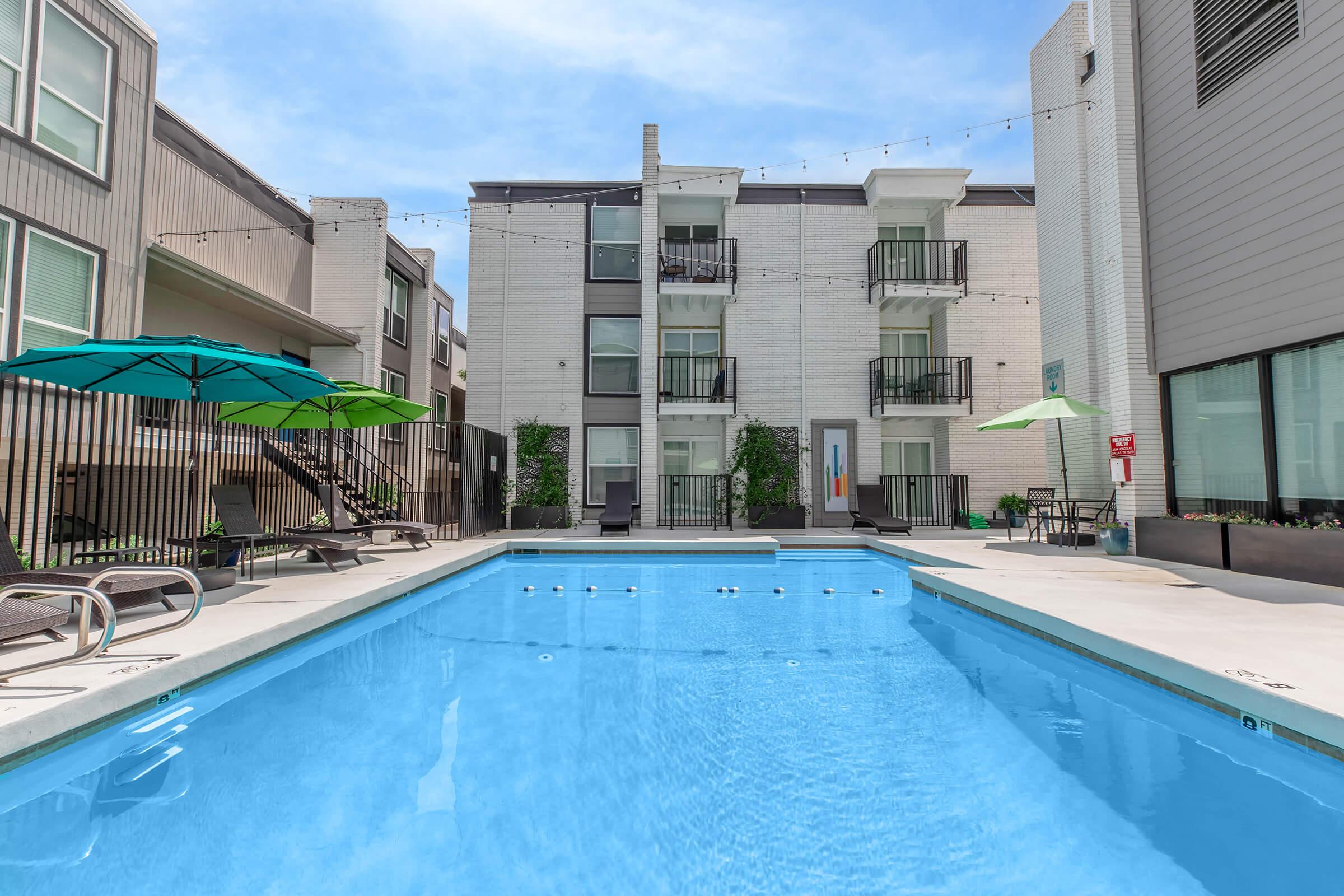 A clear swimming pool surrounded by lounge chairs and umbrellas in a modern apartment complex. There are two buildings visible, one with balconies and greenery. The sky is bright and blue, adding to the inviting atmosphere of the pool area.
