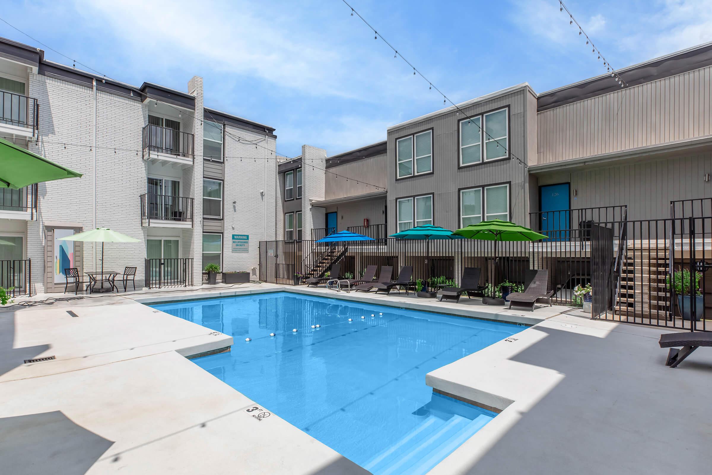 A bright outdoor swimming pool surrounded by lounge chairs and umbrellas in a landscaped courtyard. Two multi-story apartment buildings are in the background, featuring balconies and a modern design. The sky is clear and blue, enhancing the inviting atmosphere.