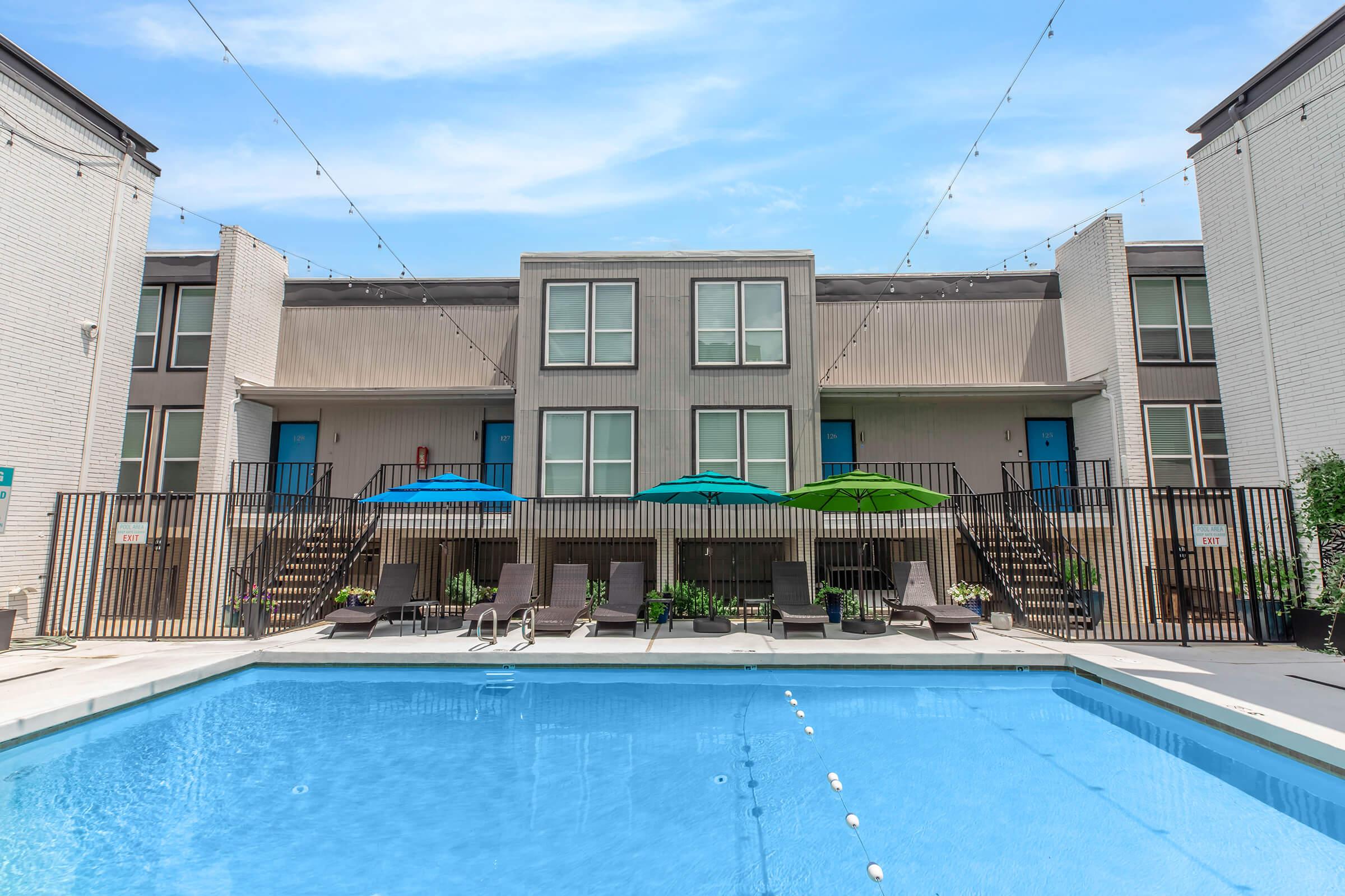 A swimming pool surrounded by lounge chairs and colorful umbrellas, with a backdrop of a two-story apartment building. The building features balconies with stairs leading down to the pool area, and the scene is set under a clear blue sky with decorative string lights overhead.