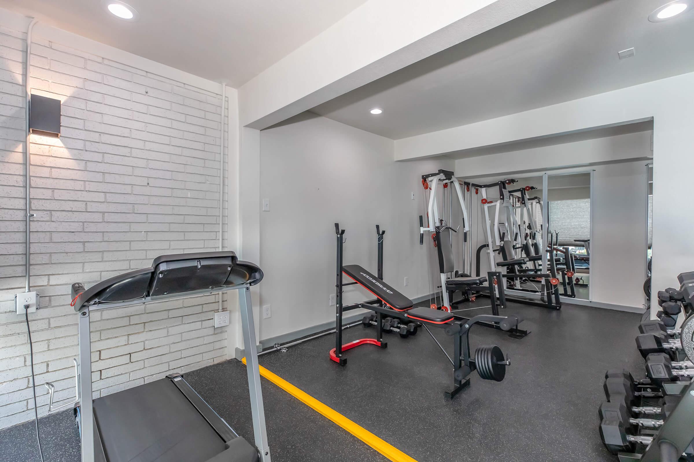 A modern home gym featuring a treadmill, weight bench, cable machines, and free weights. The space has white brick walls, gray flooring, and mirrors reflecting the equipment, creating a bright and inviting workout environment.