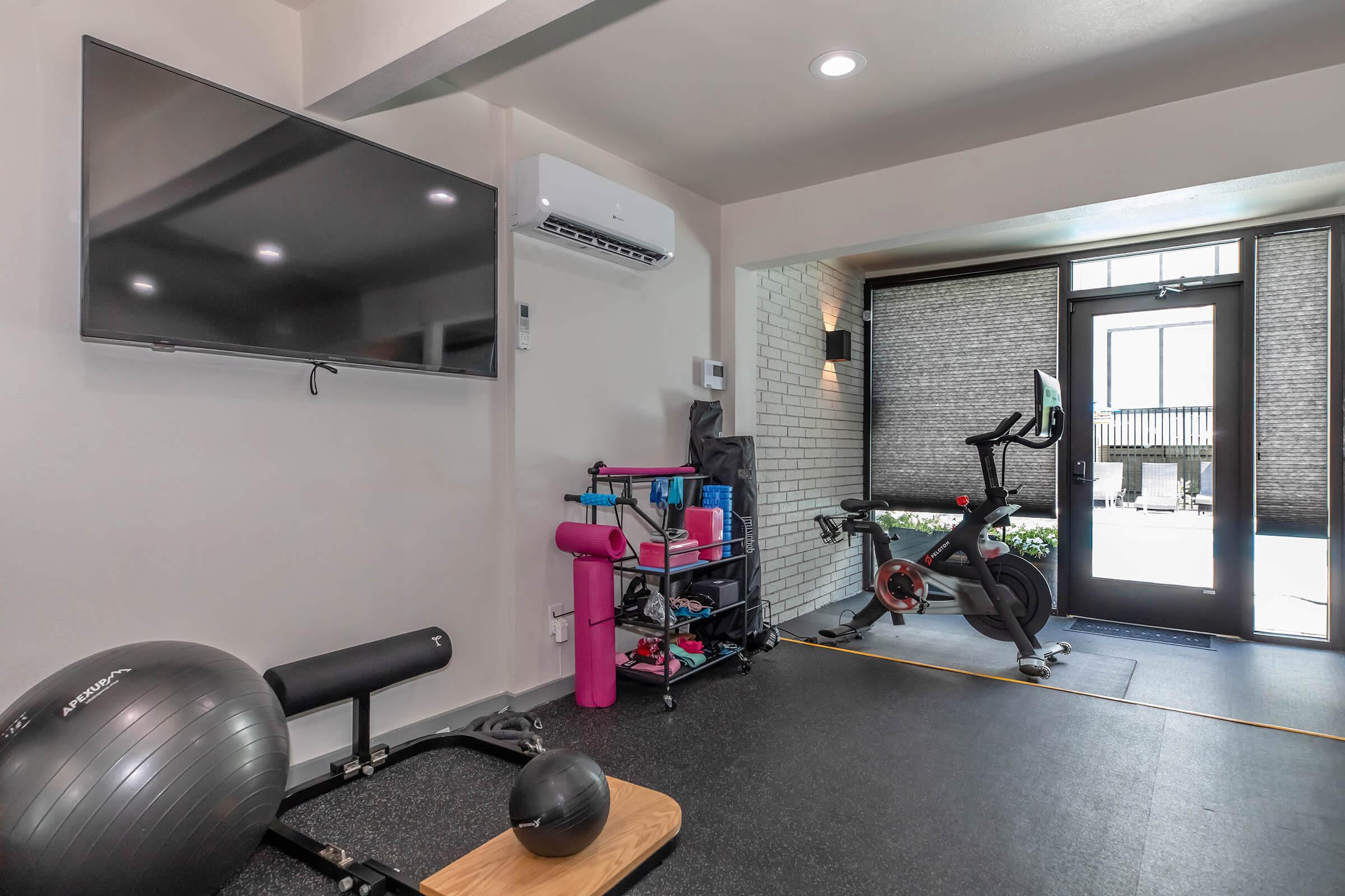 A modern home gym featuring a stationary bike, exercise mat, and a balance ball. A TV mounted on the wall and storage shelf with fitness equipment are visible. Large windows provide natural light, and a door leads to an outdoor area. The space is clean and minimalist, promoting an inviting workout environment.