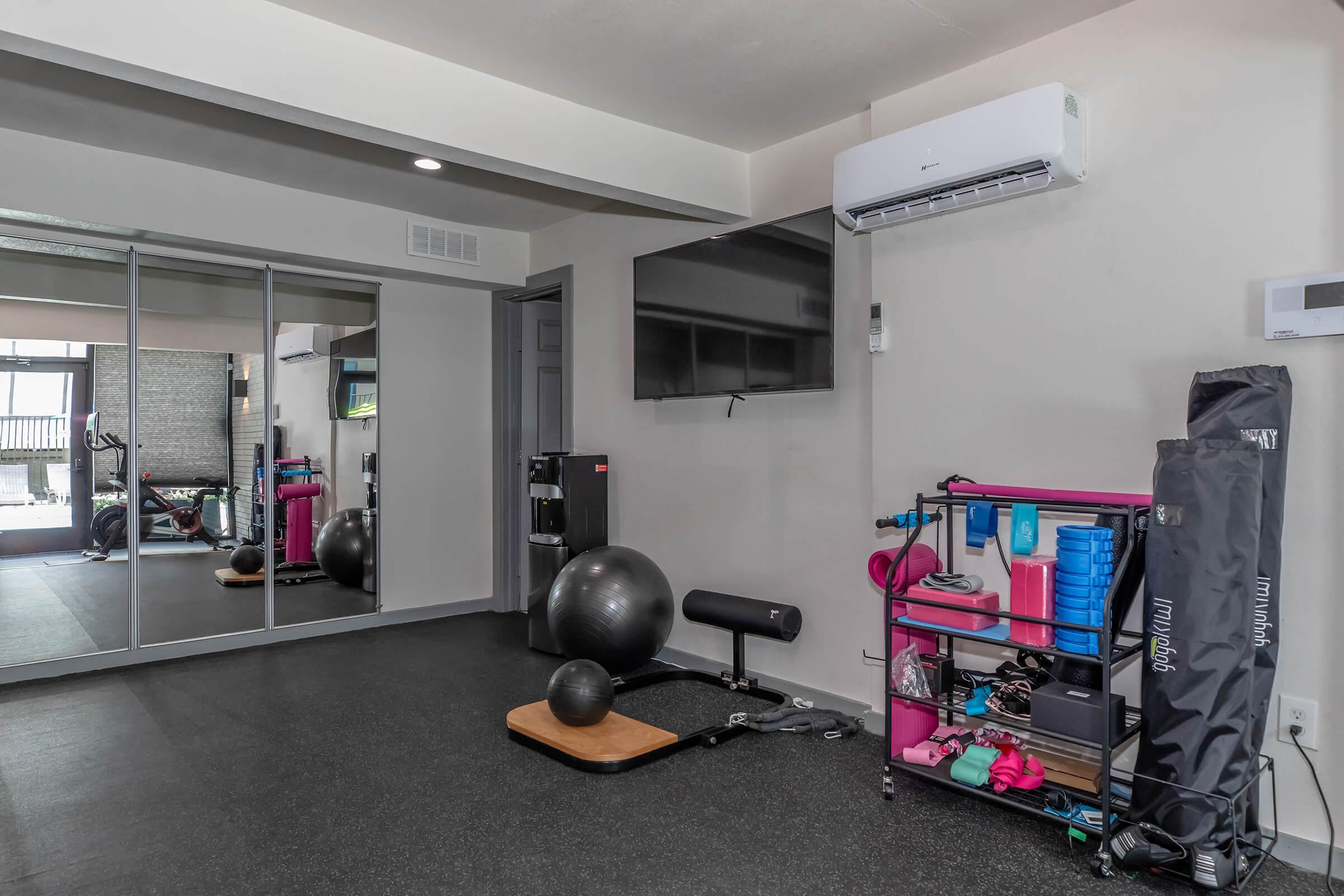 A modern home gym featuring exercise equipment including a stability ball, weights, and resistance bands on a colorful shelf. A large mirror reflects the space, enhancing light and depth. An air conditioning unit and a wall-mounted TV are also visible, creating a functional workout environment.