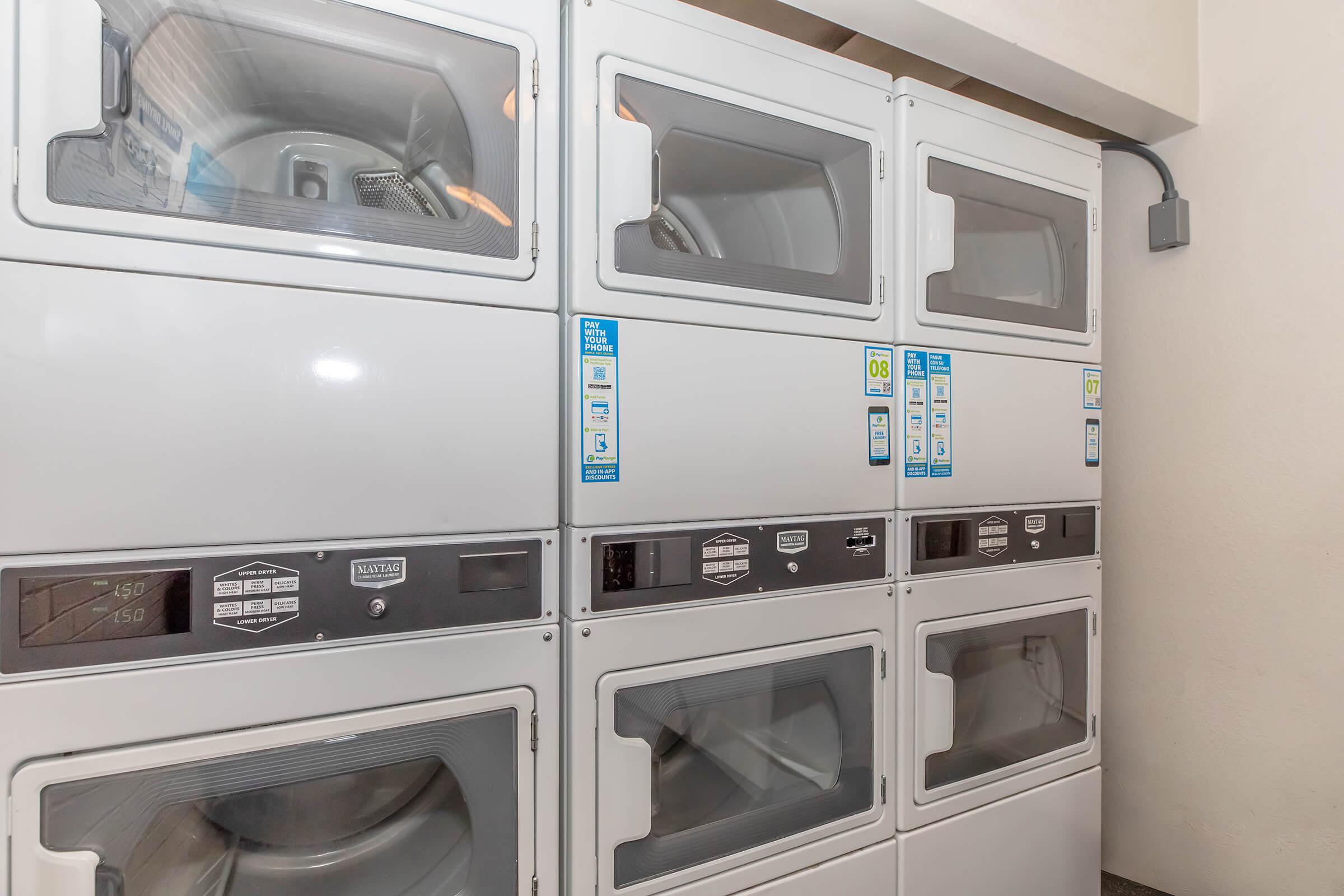 A row of stacked, modern laundromat machines for washing and drying clothes, featuring clear glass doors and various buttons and labels. The setting appears clean and functional, designed for efficient laundry use.