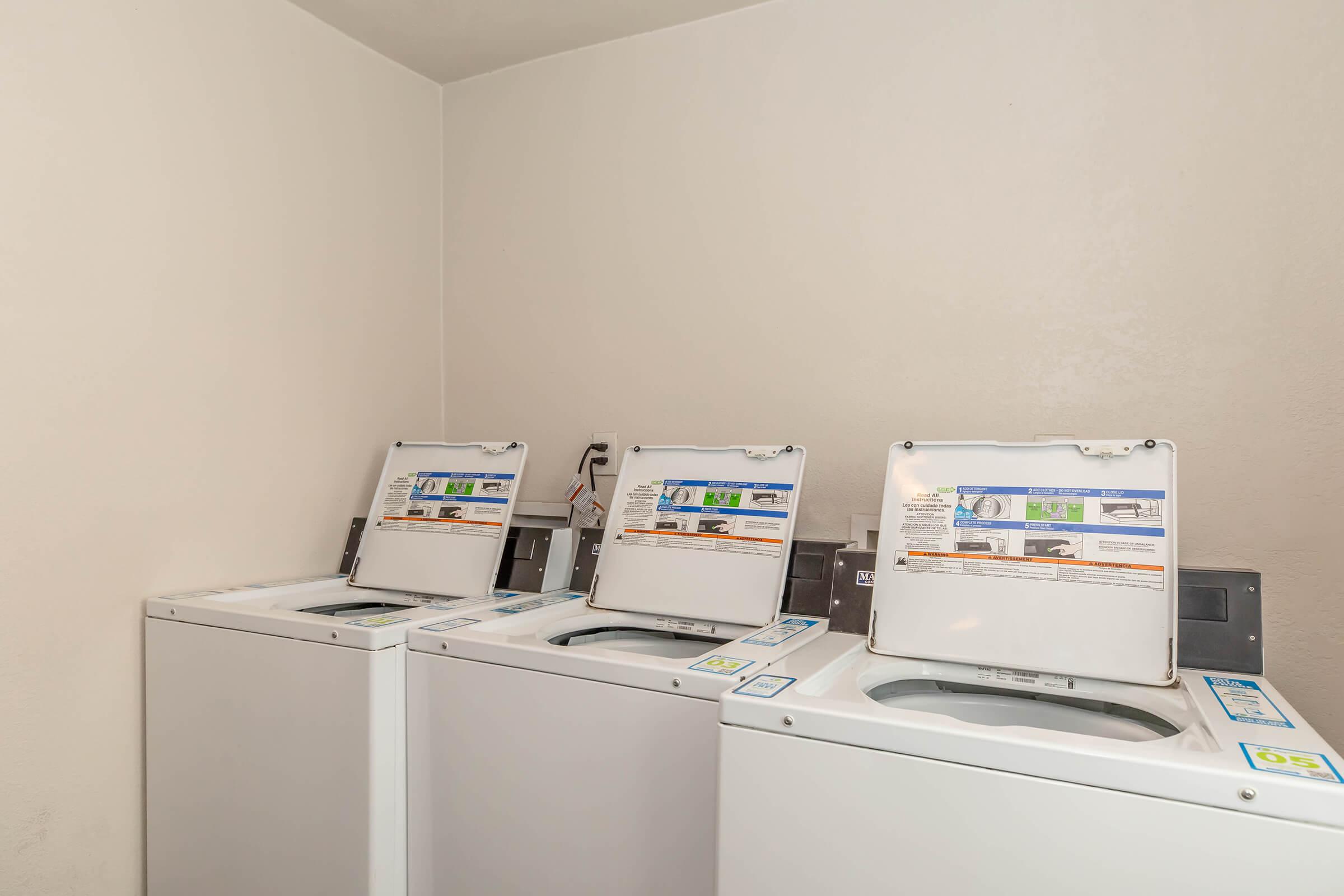 Three white washing machines lined up in a laundry room, each with a raised lid displaying controls and instructions. The walls are plain and light-colored, creating a clean and functional space for laundry activities.