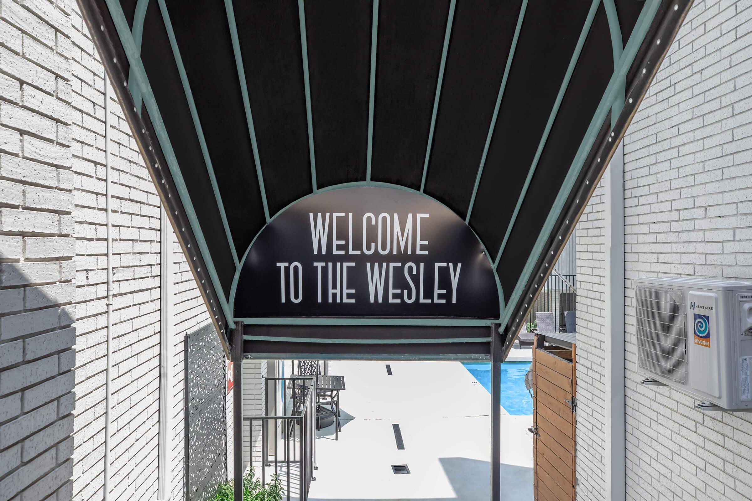 A covered entrance featuring a sign that reads "WELCOME TO THE WESLEY." The backdrop includes a clear view of a pool area, with lounge chairs visible. The architectural design has a modern aesthetic with brick walls and a sloped ceiling.