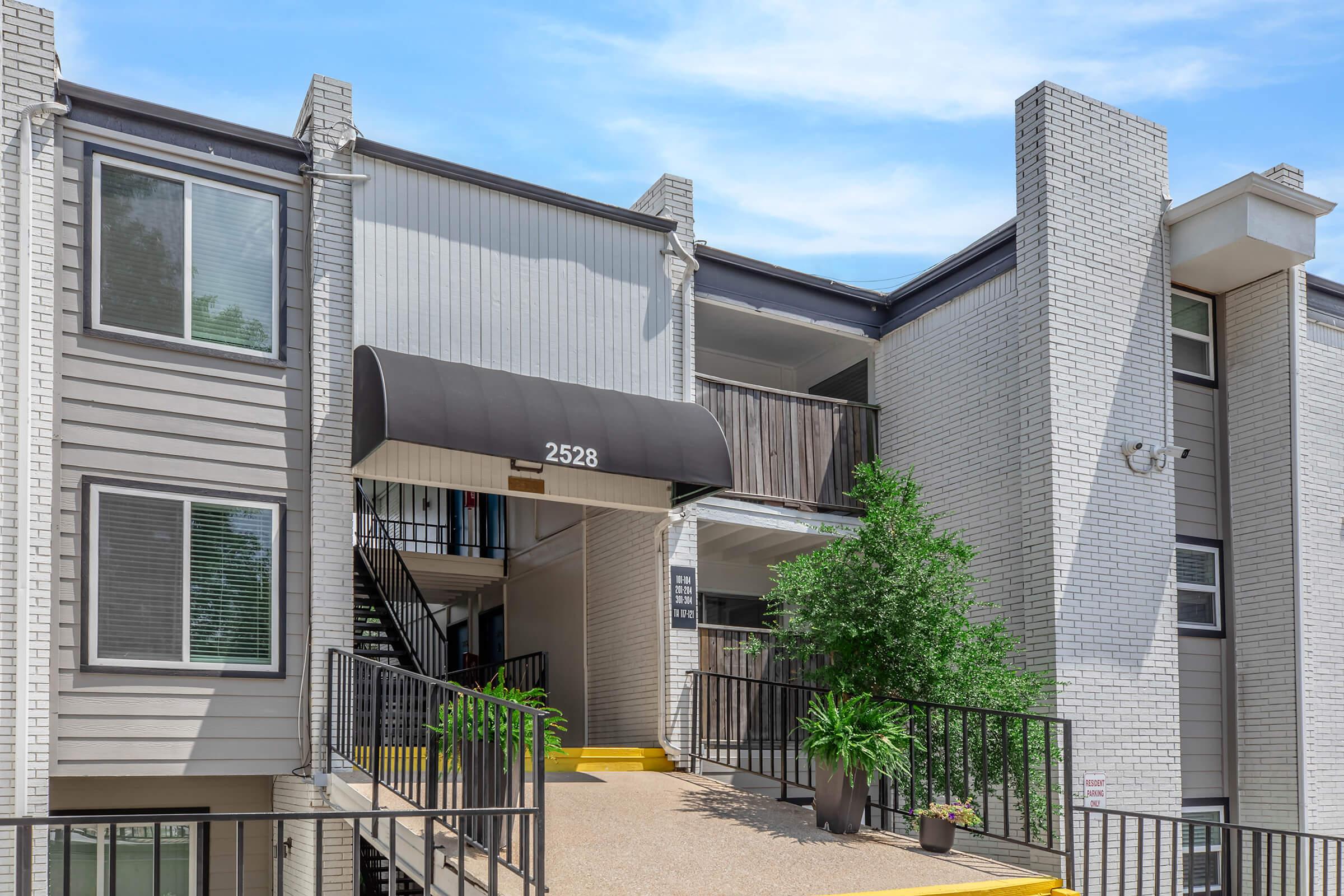 Exterior view of a multi-story apartment building featuring an entrance with a black awning labeled "2528," flanked by greenery and a staircase leading up to the second floor. The building has a mix of light and dark siding, with a clear blue sky in the background.