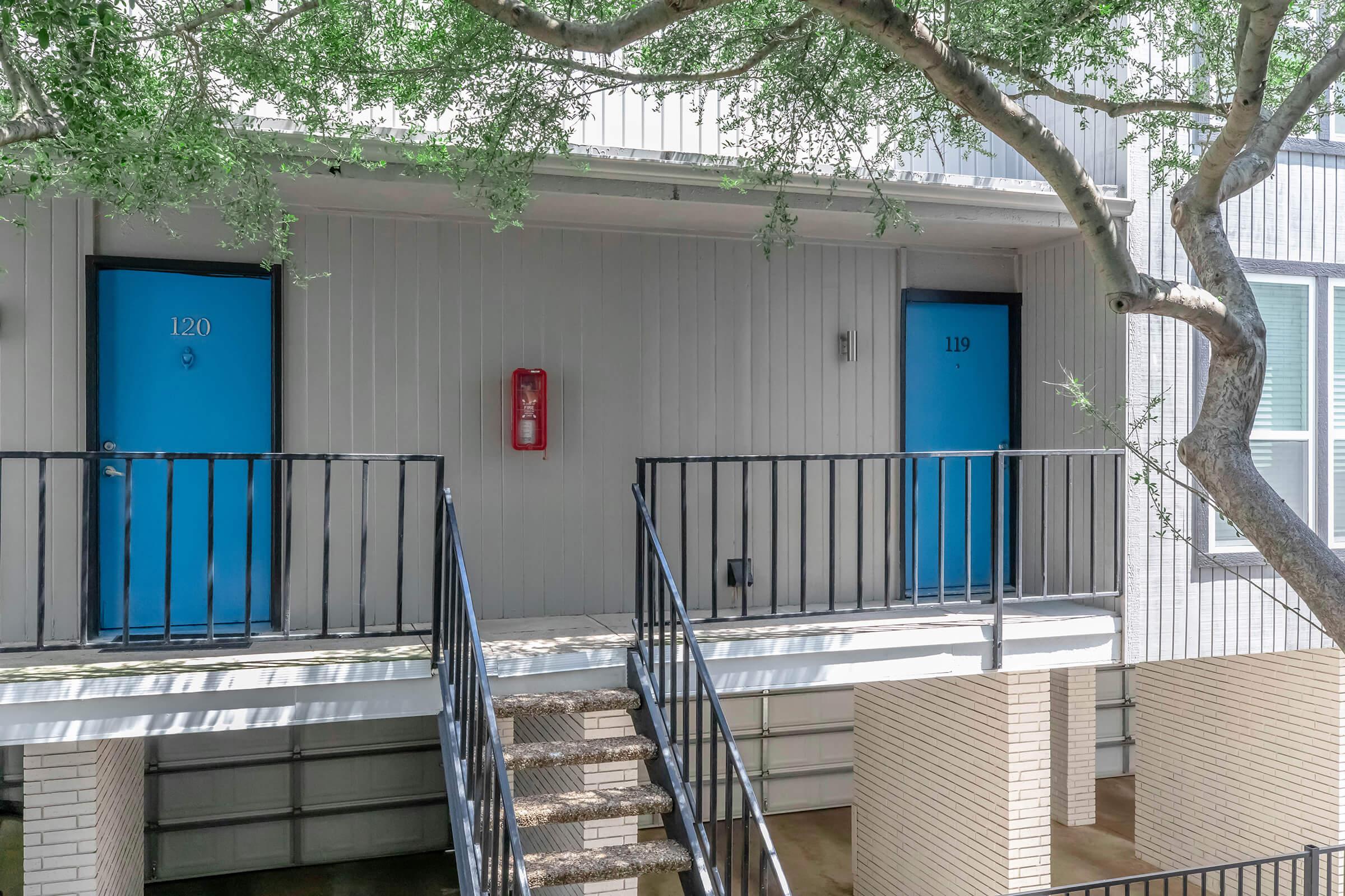 Two blue doors labeled 120 and 119 on a lightweight balcony, with a fire extinguisher mounted on the wall between them. A tree grows nearby, providing shade over the entrance area. The building has a modern architectural style, with concrete elements and a metal railing.