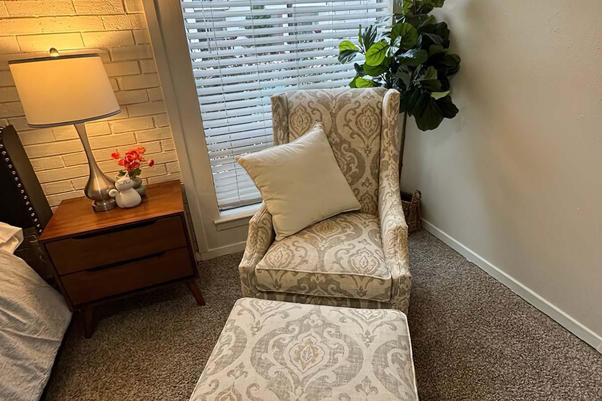 A cozy corner of a room featuring a patterned armchair with a white pillow and an ottoman. A wooden nightstand with a lamp sits beside the armchair, and there's a small vase with flowers on the table. Natural light streams in through a window with blinds, and a potted plant adds greenery to the space.