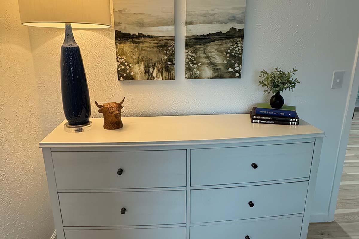 A light-colored dresser with six drawers, topped by a blue lamp, a decorative wooden bull figurine, and a small plant. Above the dresser are two framed paintings depicting a serene landscape.