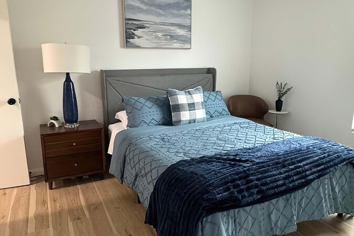 A well-decorated bedroom featuring a queen-sized bed with blue bedding and decorative pillows. There's a wooden nightstand with a small lamp and a plant. A stylish chair is positioned beside a round side table. A piece of art depicting a coastal landscape hangs on the wall, and the floor is a light-colored wood.