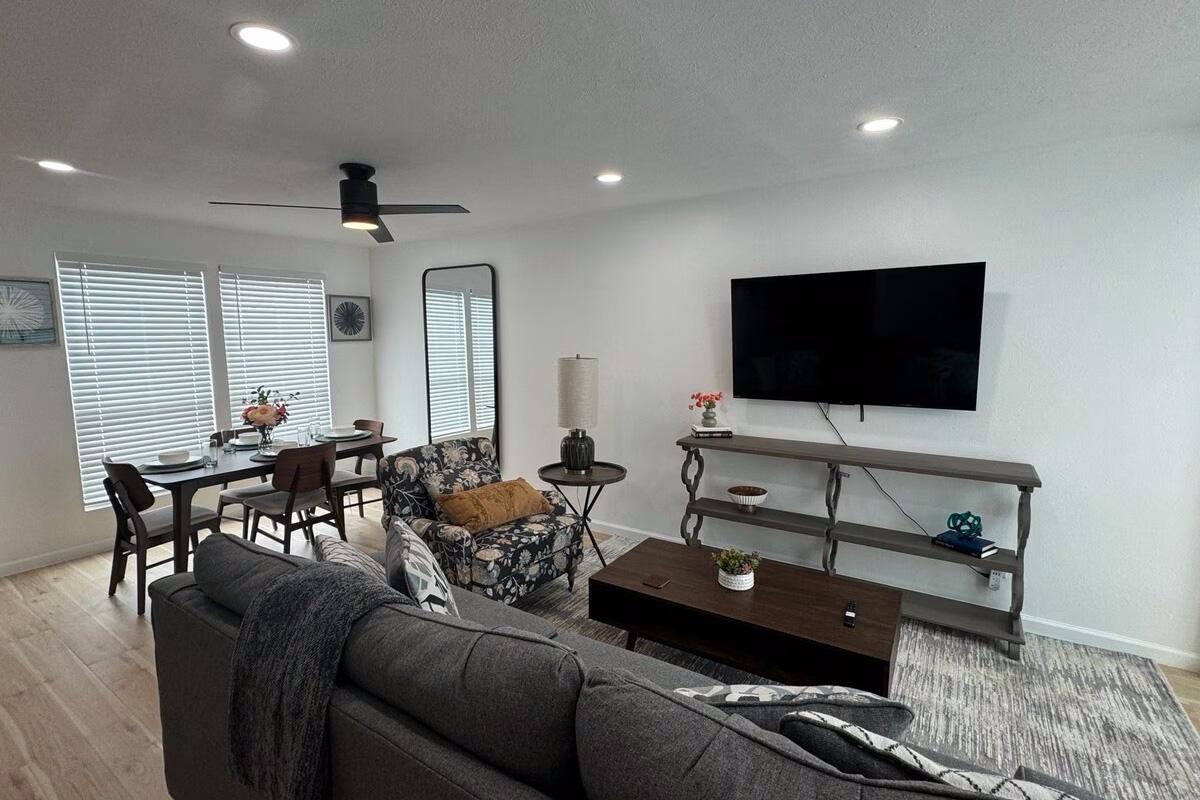 A cozy living room with a gray sofa and patterned armchair, a wooden coffee table, and a TV mounted on the wall. There's a dining area with a table set for four, surrounded by bright natural light from large windows with blinds. A floor lamp and decorative items add to the inviting atmosphere.