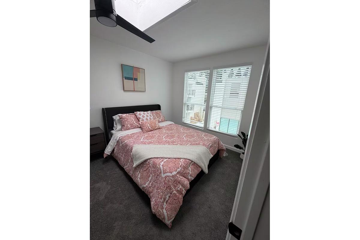 Cozy bedroom featuring a queen-sized bed with a patterned pink and white duvet, two large windows with blinds, a nightstand, and a piece of abstract art on the wall. Natural light fills the room, adding a warm and inviting atmosphere.
